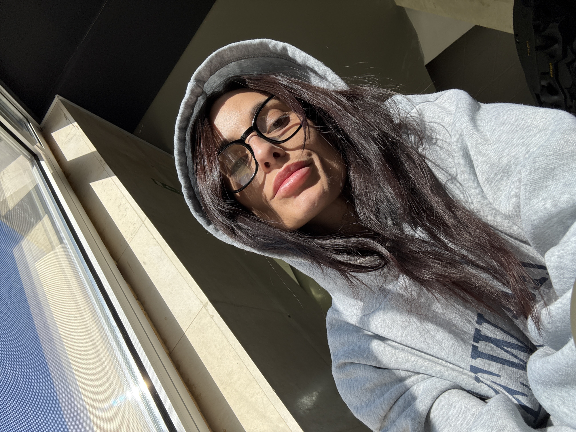 A woman with dark hair and glasses, wearing a gray hoodie, poses for a selfie near a window. The location is within a building with beige tiled walls and a window overlooking a sunny day. The exact location within Pleven, Bulgaria is indeterminable from the image.