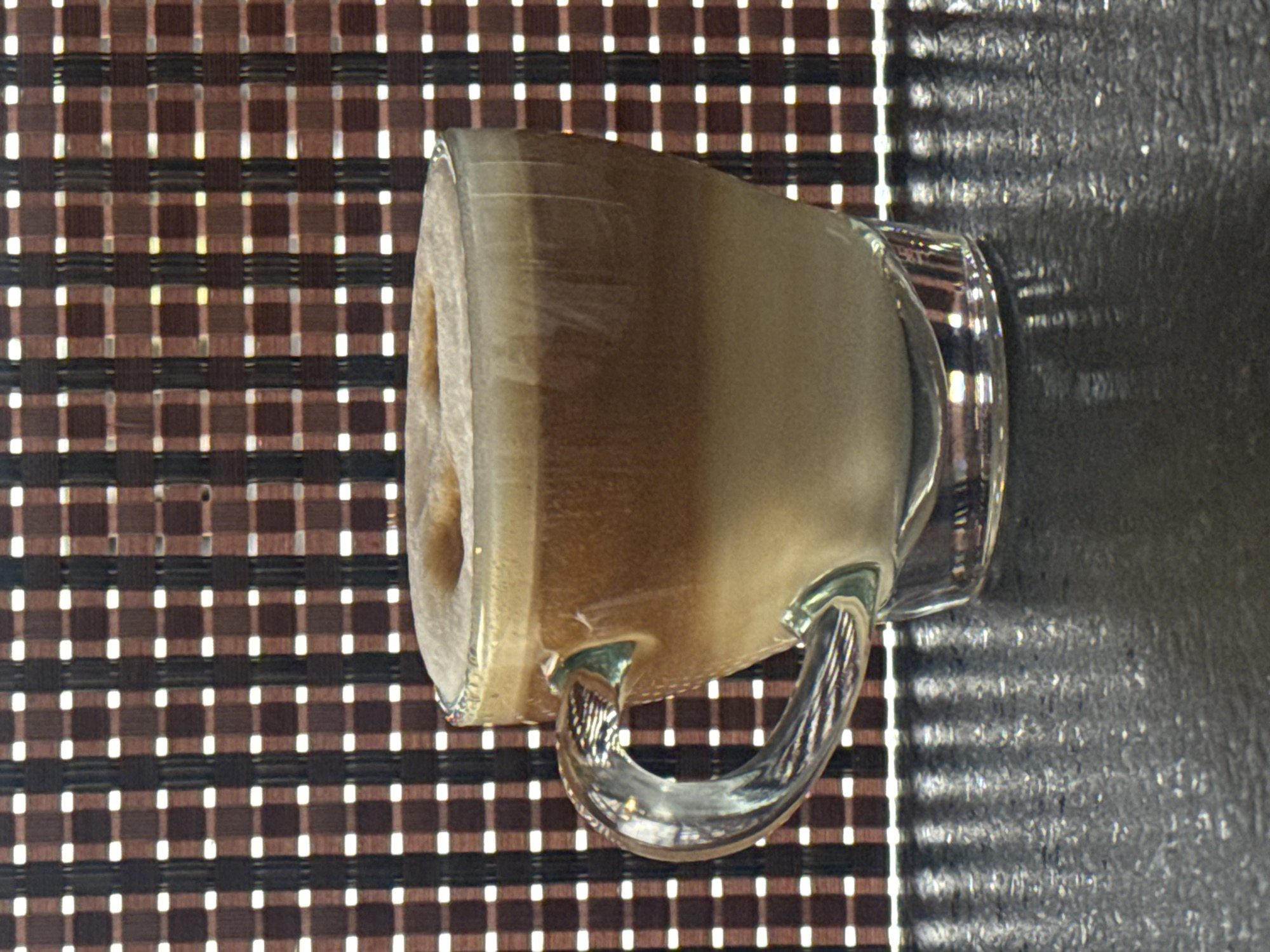 The image depicts a glass mug filled with a light brown beverage, likely coffee or a similar drink. The mug sits on a surface covered with a woven brown and white checkered pattern, resembling a placemat. To the right of the mug is a dark, textured surface, possibly a countertop. The lighting suggests an indoor environment with daylight entering from the right, casting a shadow of the mug onto the dark surface. There are no people visible in the image. The city and location are unknown.