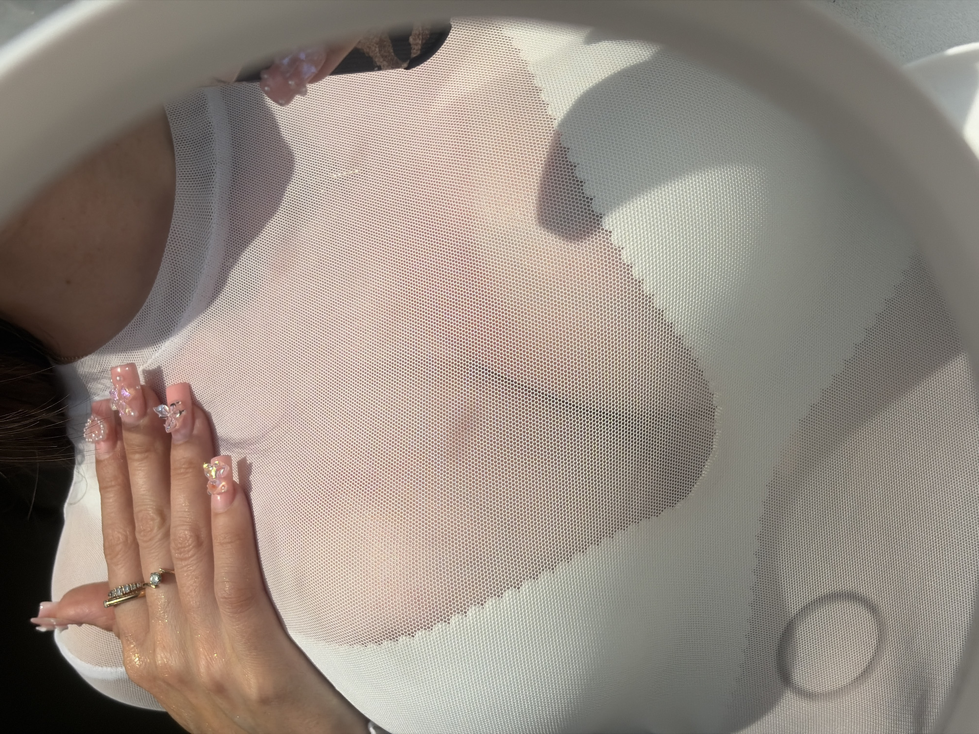 The image is a close-up shot of a person's chest and hand, wearing a sheer white mesh top over a white undergarment. The person has long, acrylic nails with elaborate decorations, including what appear to be small gems and charms. They are also wearing a ring on their finger. The background is blurred and out of focus, suggesting an indoor setting or controlled lighting conditions, and does not provide any specific details about the city of Saint Petersburg, Russia.