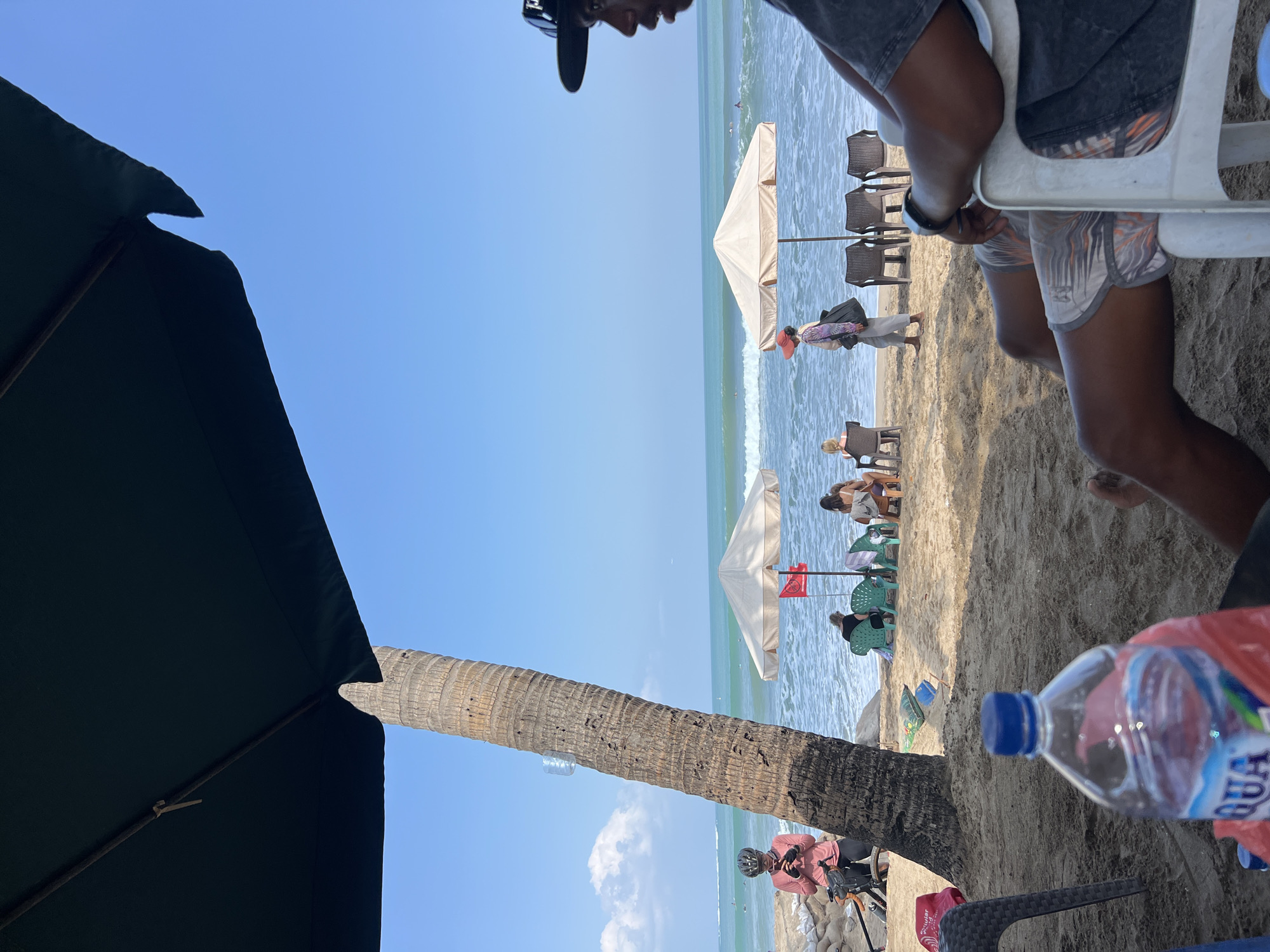 A sunny day at Kuta Beach, Indonesia, presents a relaxed scene under a large dark green umbrella. In the foreground, a person in a grey t-shirt and patterned shorts is seated on a white plastic chair, facing to the right, with their legs visible. Beside them, a bottle of AQUA water is placed on the sand, next to a red plastic bag and another chair.
Further back, a thick palm tree trunk stands prominently, leaning towards the left. A person wearing a pink long-sleeved shirt and a helmet is standing next to the palm tree, looking down at their phone, with a bicycle beside them. The beach sand is light brown, with scattered beach chairs and umbrellas in the middle ground.
Two beige umbrellas provide shade for several seated individuals. One umbrella has a red flag planted nearby. The ocean is a vibrant turquoise with moderate waves breaking on the shore. In the distance, several people are visible in the water, possibly swimming or surfing. The sky is a clear, bright blue with a few scattered white clouds. The overall atmosphere is one of leisure and enjoyment of the beach environment.