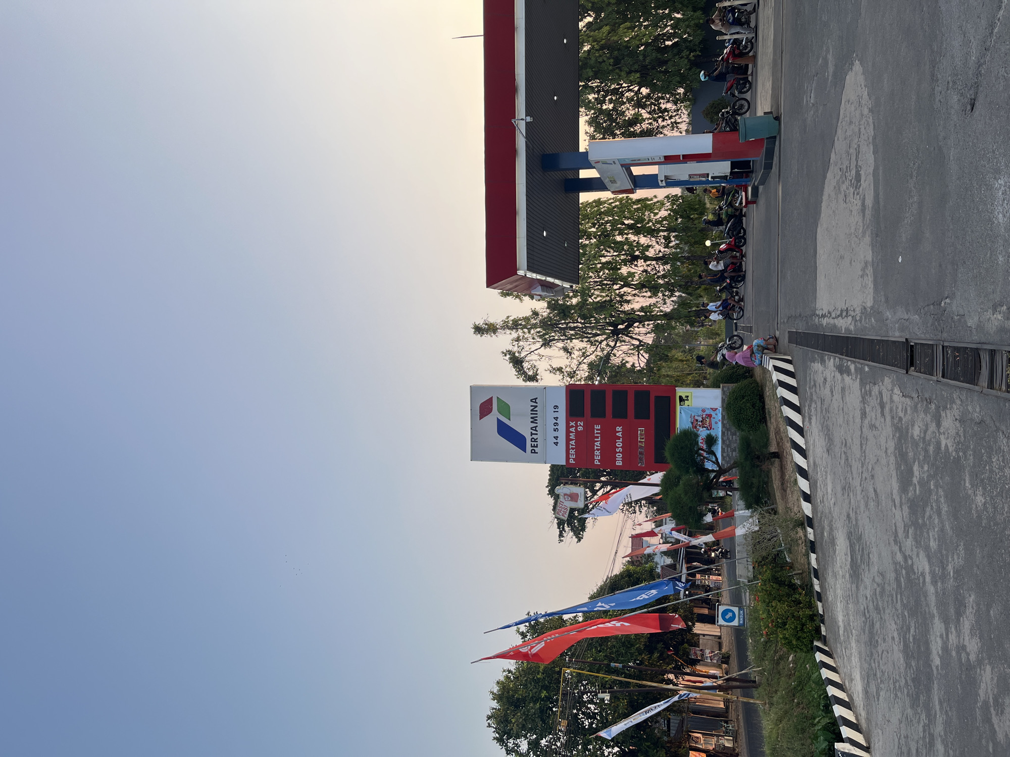 An outdoor scene at a Pertamina gas station in Pecangaan, Indonesia, captures the late afternoon light. A prominent Pertamina sign displays fuel prices, with "PERTAMAX 92," "PERTALITE," and "BIO SOLAR" listed. Several Indonesian flags, red and white, are visible. In the background, a red and white canopy covers multiple gas pumps. A group of people on motorcycles are either waiting or refueling. One individual is seated on a low concrete barrier in the foreground. The ground is paved with asphalt, and a drain runs parallel to the barrier. Trees and foliage surround the gas station, and the sky is a pale, gradient blue, transitioning to a lighter hue near the horizon, indicating the approaching sunset.