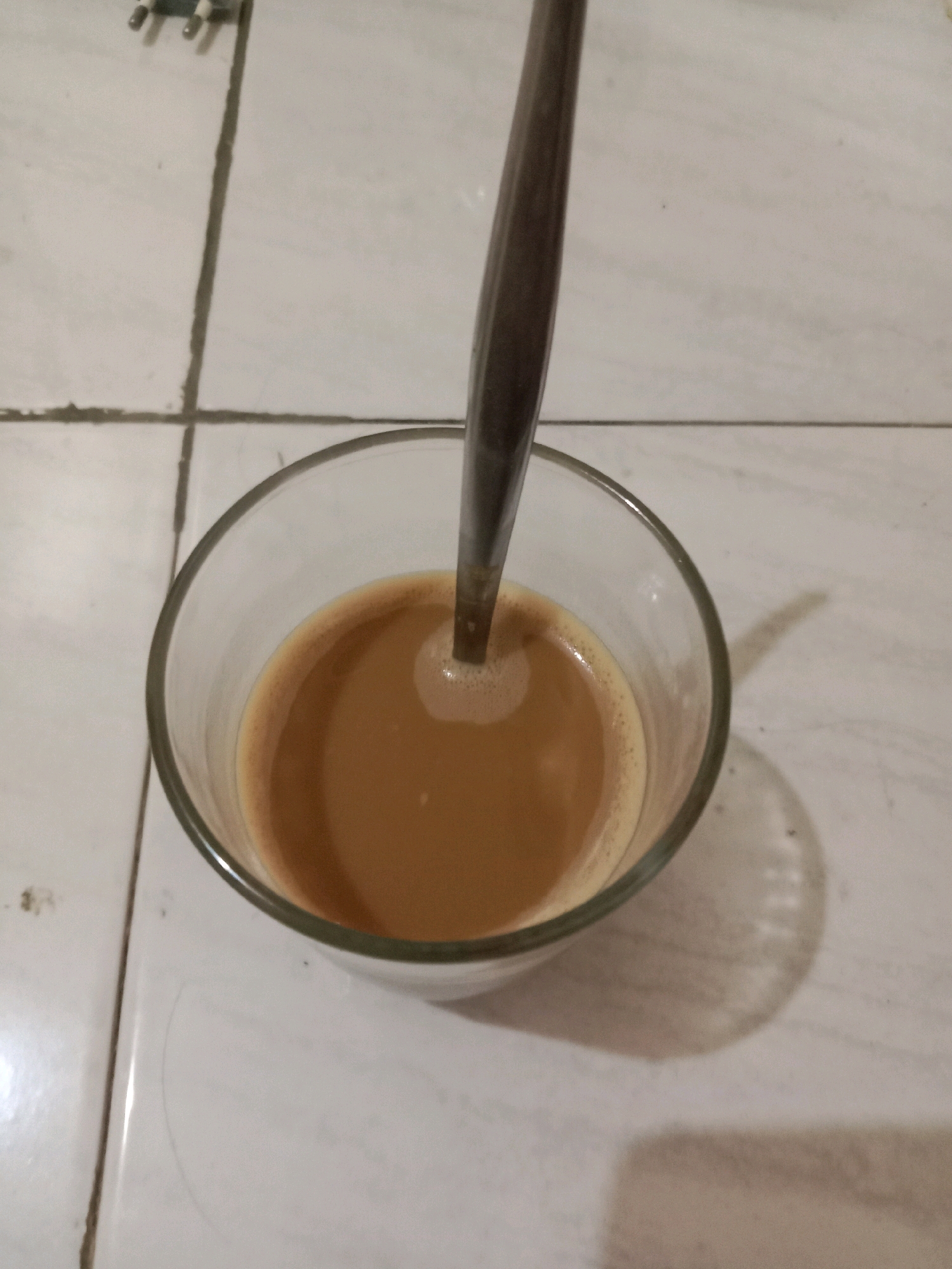 An overhead shot captures a clear glass of coffee with a spoon submerged in it. The coffee is a light brown color with a frothy surface, indicating it may have been recently stirred. The glass sits on a tiled surface, with grout lines visible in the background. The lighting suggests an indoor setting, possibly a home or a cafe. The specific location in Brangsong, Indonesia, is not discernible from the visual information alone. The image is static and shows no signs of activity beyond the presence of the coffee and spoon.