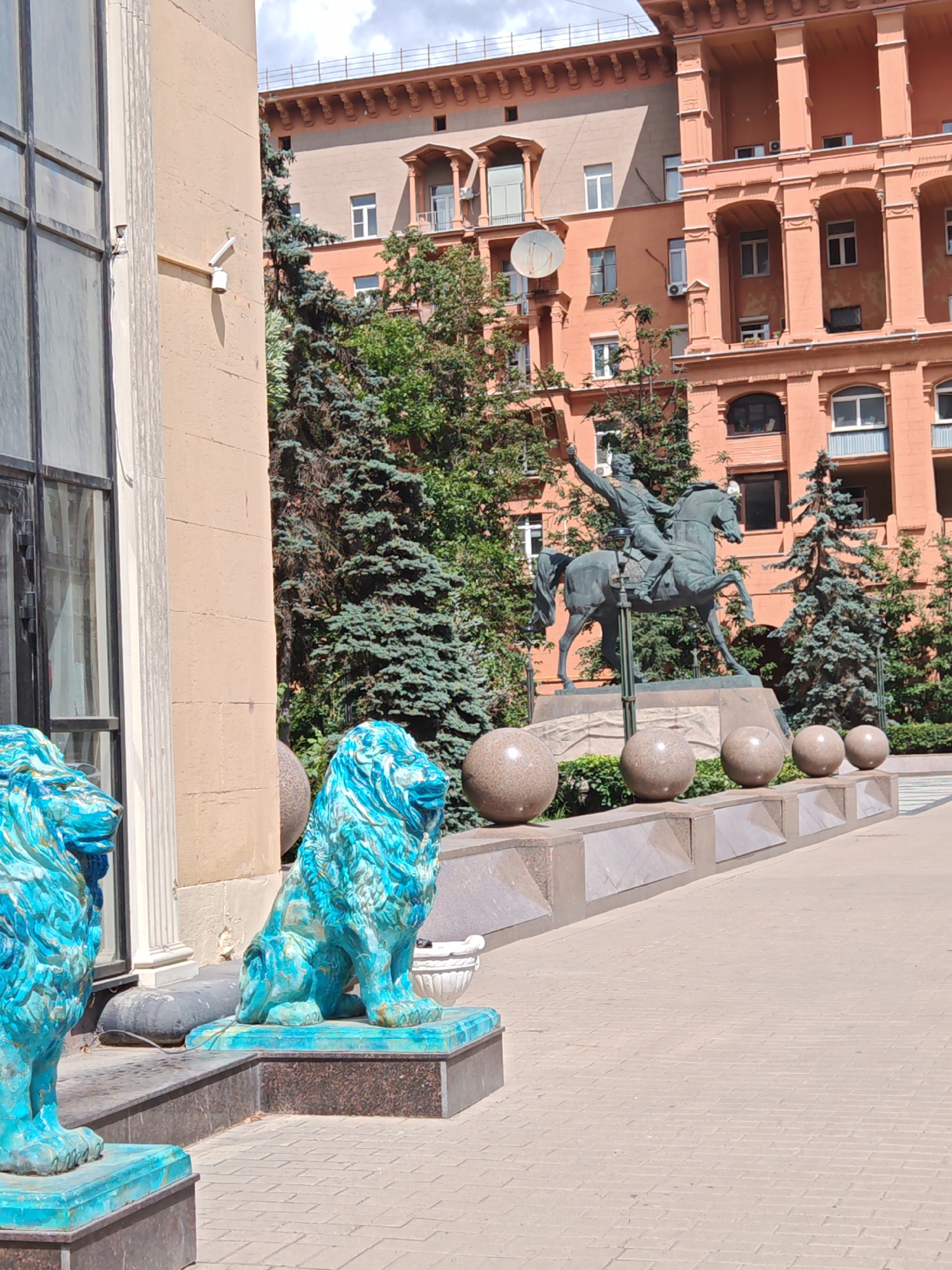 The image shows an outdoor scene in Moscow, Russia, featuring a bronze equestrian statue of a man in military uniform, with his right arm raised. The statue is positioned on a pedestal, surrounded by several large, spherical stone objects. In the foreground, two turquoise lion statues are visible, one standing and one sitting. Behind the statue and lions, there are tall evergreen trees and a large, ornate building with columns and arched windows, characteristic of Stalinist architecture, common in Moscow. The building is painted a warm, peachy-orange color. The ground is paved with light-colored bricks. The sky is visible, partly covered by clouds. The overall impression is of a public monument or square.