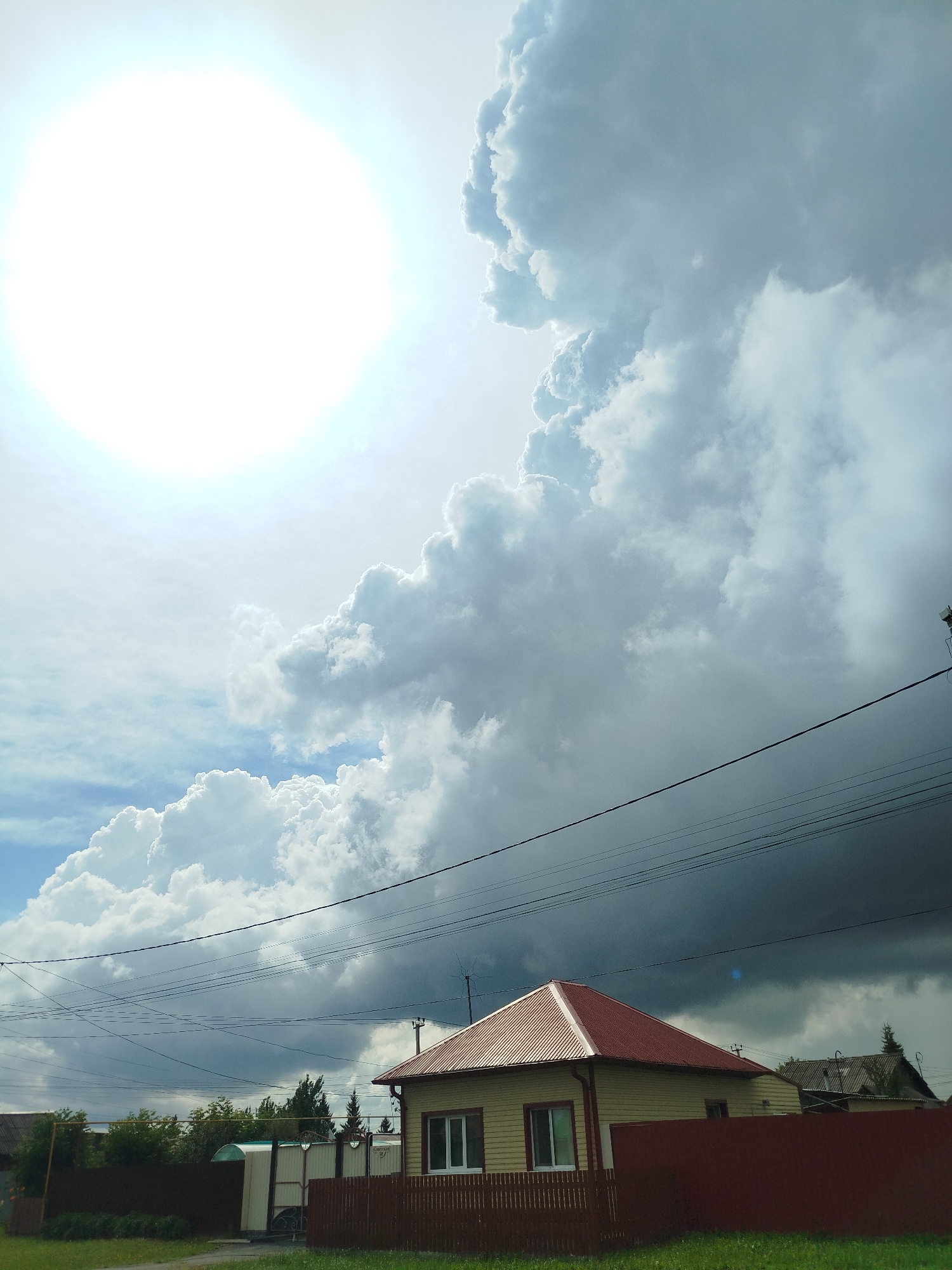 A low-angle, vertical, full shot captures a dramatic sky over a residential area in Zavodoukovsk, Russia. The upper left of the frame is dominated by a bright, almost blown-out sun, casting a strong glare. To the right of the sun, massive, textured cumulus clouds are building, with lighter white clouds at the top and darker, heavier storm clouds gathering at the bottom. A single residential building with a red, corrugated metal roof and light yellow siding is visible in the lower center of the image. A dark red wooden fence runs in front of the house and extends to the left, where it encloses a green canopy structure and a white gate. A line of trees is visible behind the fence. Multiple black power lines crisscross the sky horizontally across the midsection of the image. The ground in the foreground is covered in green grass.