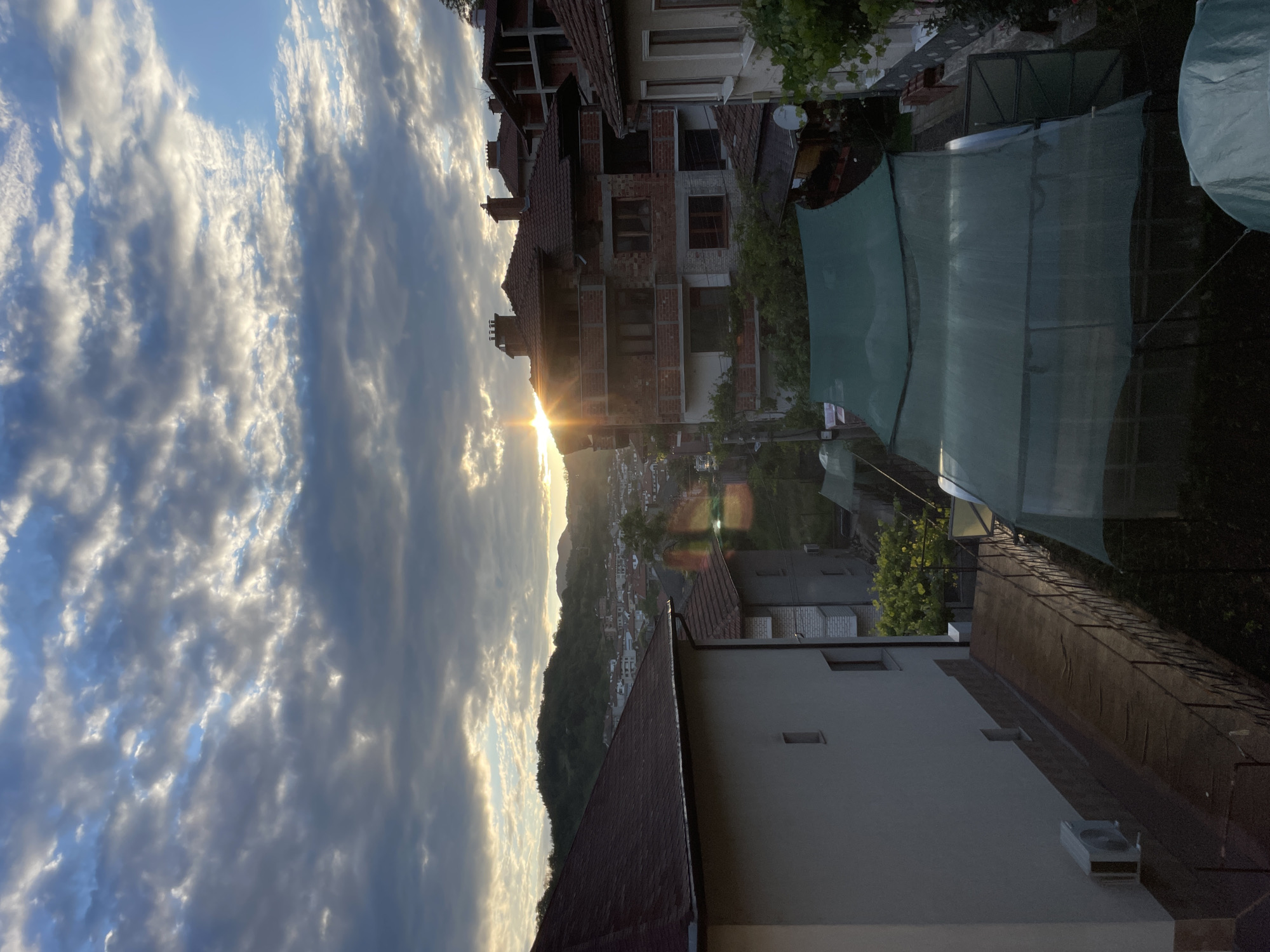 This image presents an elevated view of the city of Zlatograd, Bulgaria, during late afternoon or sunset. The sky features scattered cumulus clouds, illuminated from behind by the low sun, creating bright highlights and darker cloud formations. Patches of clear blue sky are visible, particularly in the upper right quadrant.
In the foreground on the left, the upper portion and roof of a light-colored building are visible, featuring a reddish-brown tiled roof and an external air conditioning unit mounted on a lower wall. Towards the center-right foreground, a multi-story building, appearing to be under construction or renovation, showcases exposed red brickwork, multiple balconies, and a dark tiled roof with several chimneys. Adjacent to it on the right is another building with a lighter facade and a tiled roof.
Below these structures, a garden or yard area is visible, containing several large, green netting structures, likely greenhouses or shade covers, along with a fence-like barrier running beside a narrow pathway.
The midground expands into a dense cluster of buildings, typical of a town situated in a valley, interspersed with trees and various vegetation. In the background, rolling hills covered in dense forest define the horizon. The setting sun is visible on the right, just above these hills, emitting a strong burst of light and lens flare across the scene. No individuals are present in the image.
