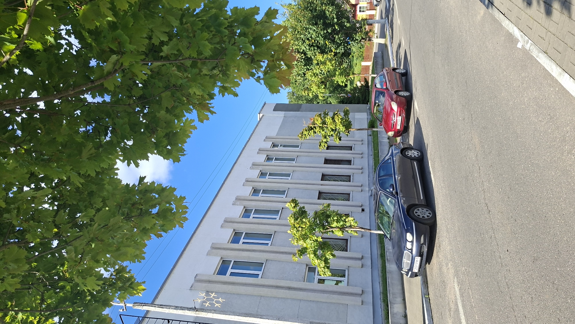 The image depicts a street scene in Myadzyel, Belarus, on a sunny day. In the foreground, a paved road is visible, with a pedestrian walkway to the right. Two cars are parked on the side of the road. A dark grey sedan, a Rover 75, is parked closest to the viewer, facing left. To its right, a red hatchback, a Citroën C2, is also parked, facing left.
Behind the cars, a light-colored, multi-story building with a row of windows and vertical concrete elements runs along the street. Two small trees with green foliage are planted in front of the building. Overhead, the branches of a large tree with green leaves obscure a portion of the sky, which is bright blue with a few white clouds. In the background, more trees and a glimpse of houses suggest a residential or mixed-use area. The overall impression is of a calm, urban environment.