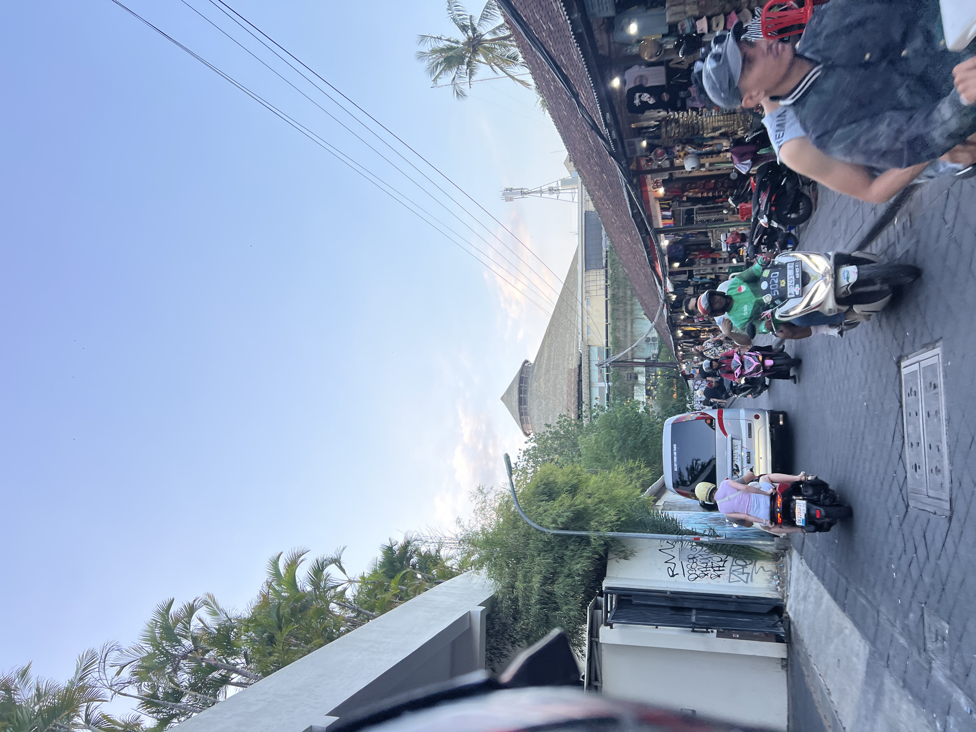 In Kuta, Indonesia, a street scene depicts a busy thoroughfare lined with shops and stalls. The time of day appears to be late afternoon or early evening, with soft, diffused light. A person in a green shirt with "Grab" branding is riding a white scooter, followed by a person in a grey jacket wearing a baseball cap. To their left, a white van is parked, with graffiti on the adjacent wall. Several other scooters and motorcycles are also visible on the street, with a mix of riders and passengers. In the background, a building with a distinctive thatched roof structure is visible, along with palm trees and power lines stretching across the sky. The shops on the right side of the street display various merchandise, with some lights illuminating the goods. The street is paved with interlocking stones, and drainage grates are present.