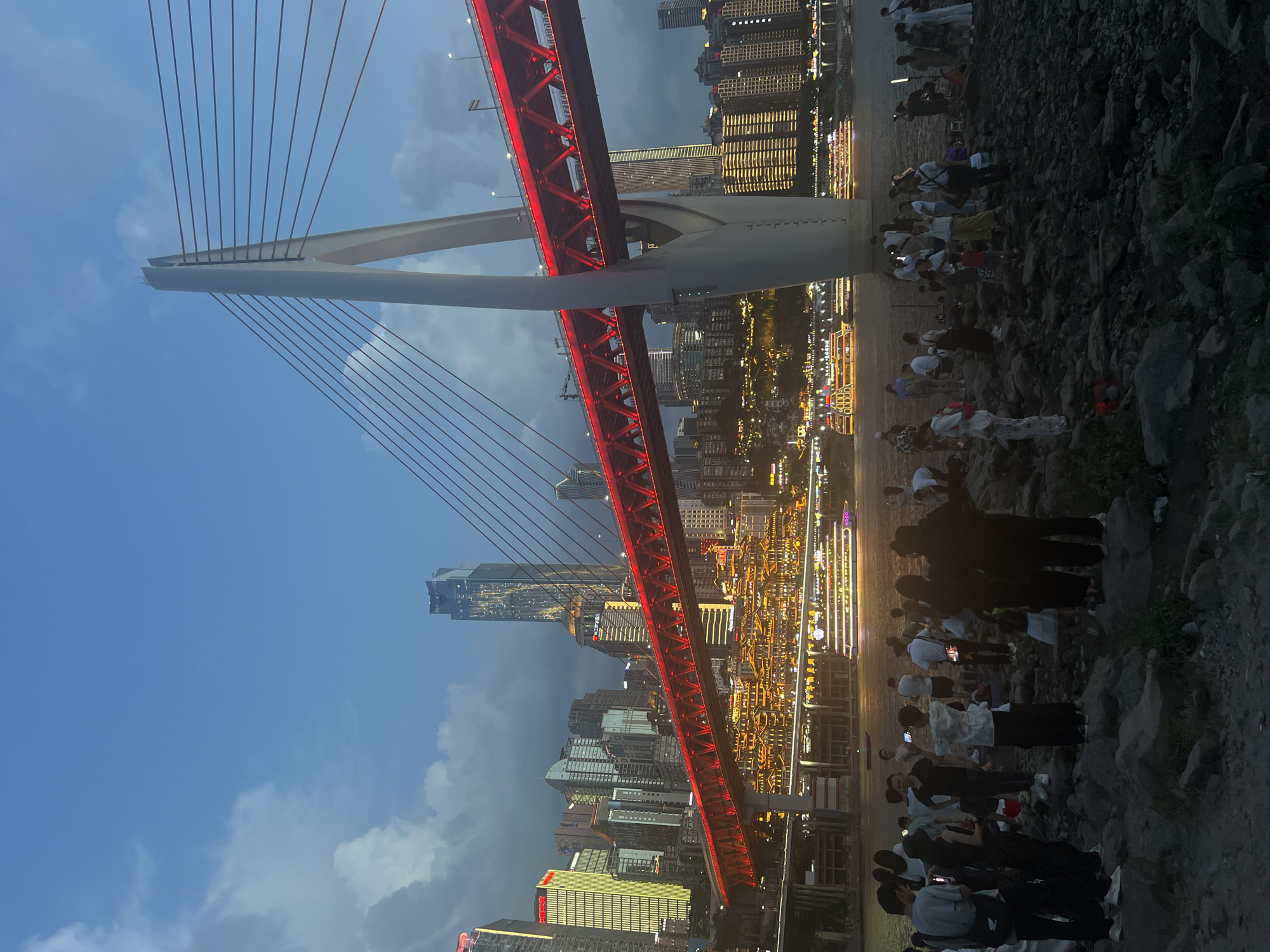 The image depicts a night scene along the waterfront of the Yuzhong District in China. A modern cable-stayed bridge with its structure illuminated in red dominates the mid-ground, spanning a wide river. Across the river, a dense collection of skyscrapers and buildings, many lit with yellow and white lights, form the city skyline. The lower portions of the buildings on the riverbank are also illuminated, creating a festive atmosphere. Several tour boats with bright lights are navigating the water. In the foreground, a rocky embankment is populated by a crowd of people. They are standing, sitting, and interacting, many holding up phones, suggesting they are observing the illuminated bridge and cityscape or possibly taking photographs. The sky is a deep blue, with some scattered clouds visible, and the overall lighting indicates it is dusk or early evening. The scene conveys a lively urban environment at night.