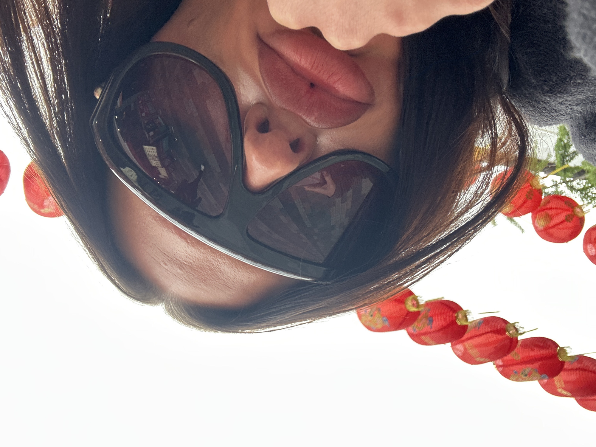 The image depicts a woman wearing large, dark sunglasses, upside down against a bright, white background. A string of red lanterns with gold detailing arcs along the bottom edge of the frame, indicating a festive or cultural event. The location is suggested by the lanterns and architectural reflections in the sunglasses to be in a Chinatown or similar district, specifically in the city of London, United Kingdom. The woman’s face, including her lips and nose, are centrally positioned. A piece of gray clothing is visible on the right.