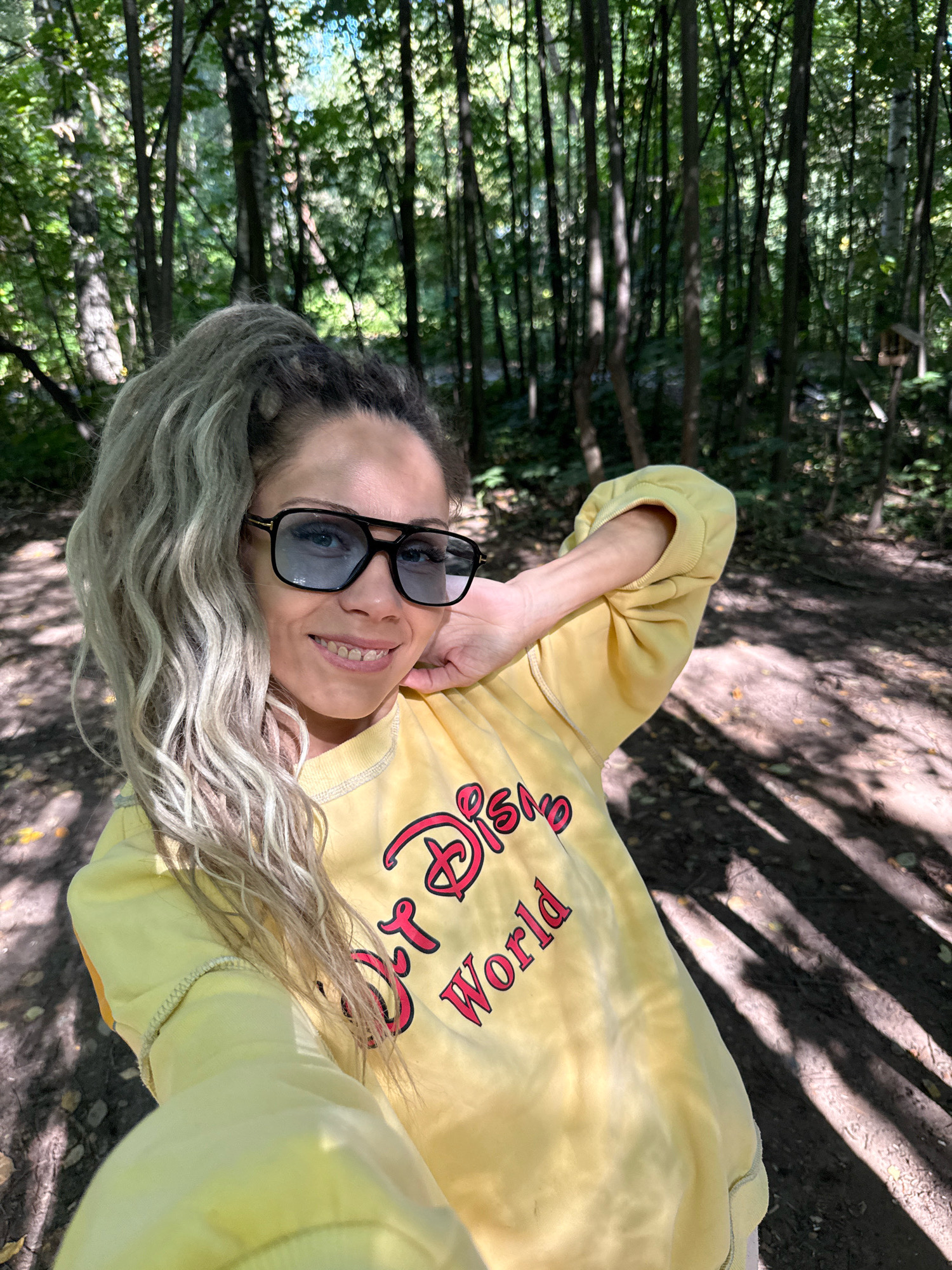 A woman is taking a selfie in a wooded area in Rusanovo, Russia. She is wearing a yellow sweatshirt with "Disney World" printed on it in red lettering. She has on sunglasses and her hair is pulled back in a high ponytail. The background is a forest with trees and dappled sunlight.