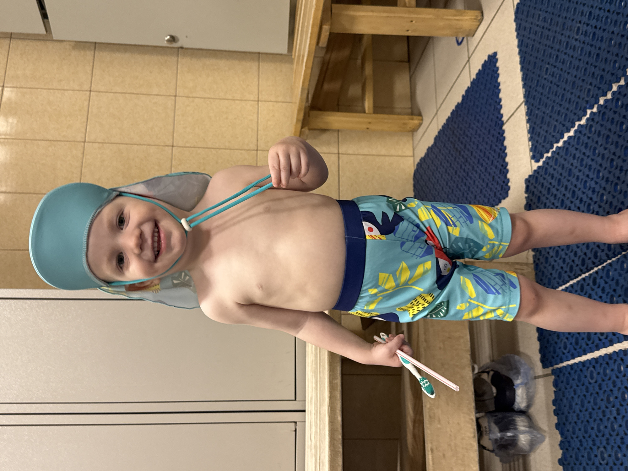 A young boy with a bright smile and a light blue swim hat, adorned with a playful shark fin and tail, is the main subject. He is wearing blue swim trunks with a tropical pattern of green leaves and yellow fruit. His right arm is extended, holding two straws, one pink and one white, as if for a drink. His left hand is holding onto the teal drawstrings of his swim hat.
The boy is standing on a blue, textured mat, which is placed over light-colored floor tiles. Behind him, a wooden bench with horizontal slats is visible, suggesting a public restroom, changing room, or poolside area. The overall lighting suggests it is daytime. The setting appears to be indoors, possibly a locker room or a changing area at a swimming facility in Saint Petersburg, Russia. The boy's expression is joyful and engaged. There is no visible text in the image.