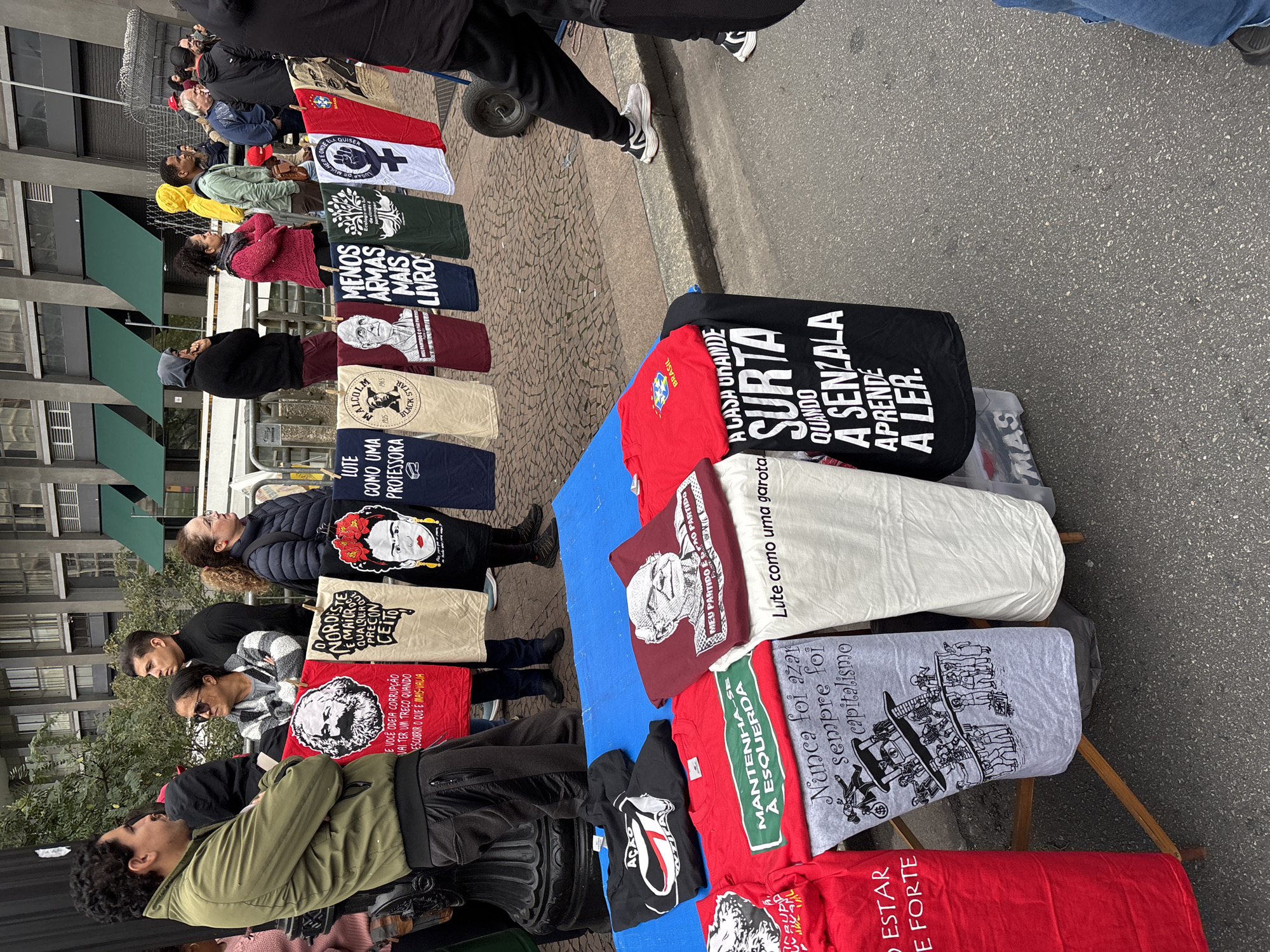 The image shows a street vendor's stall selling printed T-shirts and tote bags in what appears to be a public space. Customers are browsing through the merchandise. The T-shirts feature various political and social messages, including images of historical figures such as Frida Kahlo, Malcolm X, and Karl Marx, as well as slogans related to education, feminism, and anti-capitalism. The vendor stall is set up on the sidewalk, near a building with awnings. The location is potentially São Paulo, Brazil, due to the presence of Brazilian flags and Portuguese phrases on some of the merchandise.
