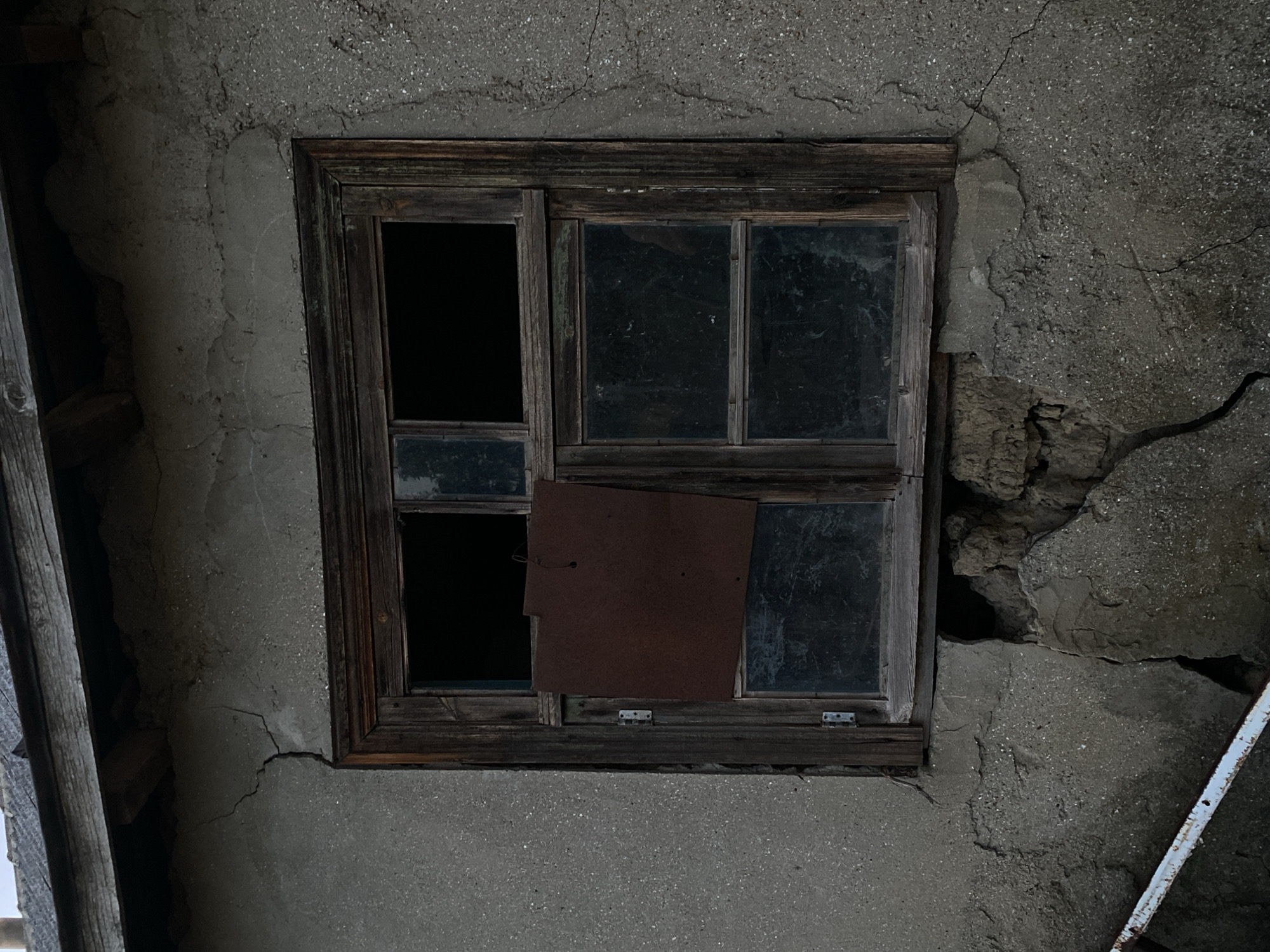 The image shows a close-up of a weathered window on the side of an old building, likely in the village of Cairaclia, Moldova. The building's facade is rough, textured concrete with visible cracks, suggesting disrepair. The wooden window frame is dark and aged, with some peeling paint or finish. The window itself is divided into panes, some of which are intact and dark with reflection, while others are open and black. A square piece of rusty metal is affixed to the inside of the window, covering a portion of one of the panes. Below the window, the concrete is significantly damaged, with large chunks missing and the underlying material exposed. A white metal object, possibly a railing or pipe, is partially visible in the bottom left corner. The overall scene evokes a sense of neglect and abandonment.