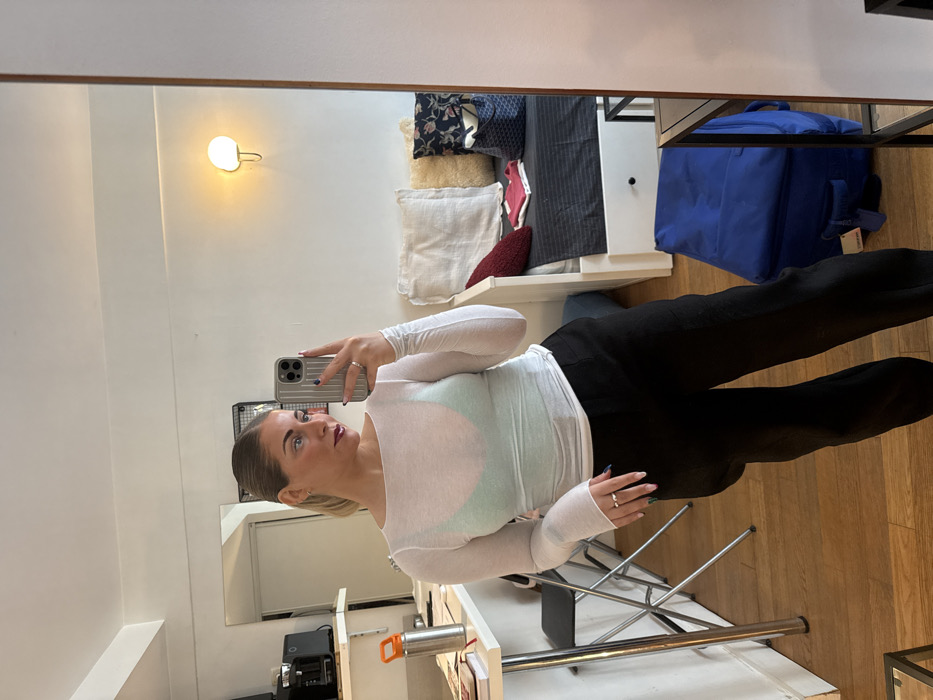 The image displays a mirror selfie taken by a female subject inside a compact residential unit in the city of Paris, France. The subject, positioned centrally, has blonde hair tied back and is wearing a sheer white long-sleeved top over a light green bralette, paired with dark trousers. She has dark red lipstick and several rings, including one on her left ring finger. Her right hand holds a silver smartphone with a textured grey case, angled towards the mirror to capture the reflection. Her fingernails are painted with various colors and patterns.
The interior features hardwood flooring and white walls. To the left of the subject, a small kitchen area is visible with a white countertop holding a silver water bottle with an orange cap and two black kitchen appliances. A metal bar stool with a black seat is tucked beneath the counter. To the right, a white daybed or single bed serves as a sofa, covered with dark striped bedding and adorned with a large cream-colored pillow, a dark patterned pillow, and a red knitted pillow. A small pink garment, a folded white item, and a navy blue patterned tote bag are resting on the bed. Above the bed, a white wall sconce with a glowing light is affixed to the wall. A blue suitcase or bag is visible on the floor in the lower right portion of the frame.