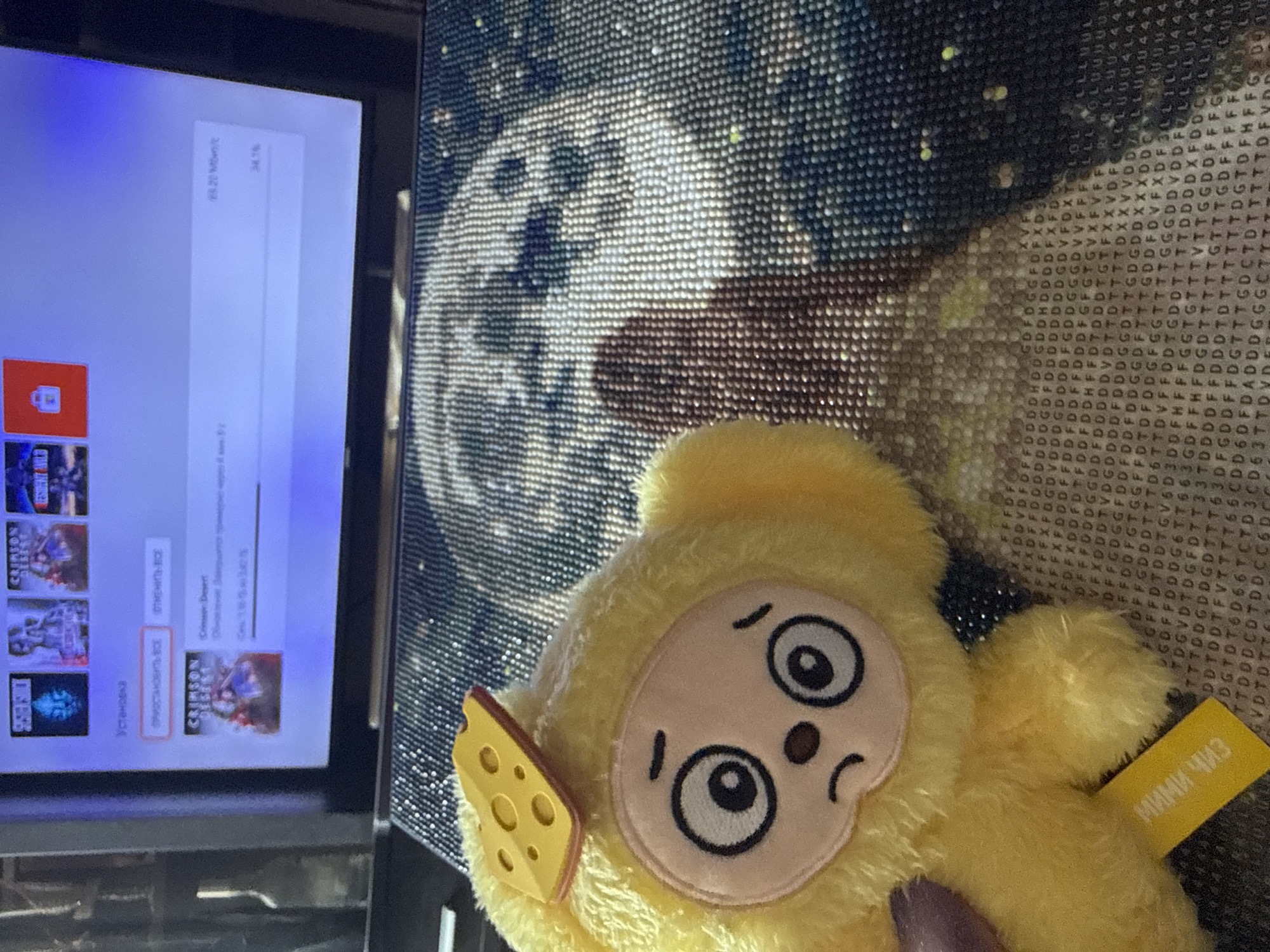 A still life scene features a television on the left displaying a video game interface in Russian, with "Crimson Desert" highlighted. To the right of the television is a diamond painting depicting an ambiguous gray and white image. Overlapping the right portion of the painting is a yellow, plush toy with a light peach-colored face, dark eyes, and a small, downturned mouth. A piece of "cheese" is attached to the left of its face and a tag labeled "МИМИ Чиз" is on the right. The scene is indoors with a dark background, and the objects appear to be positioned on a flat surface. There is no definitive information in the image to precisely establish the location in Dmitrov, Russia.