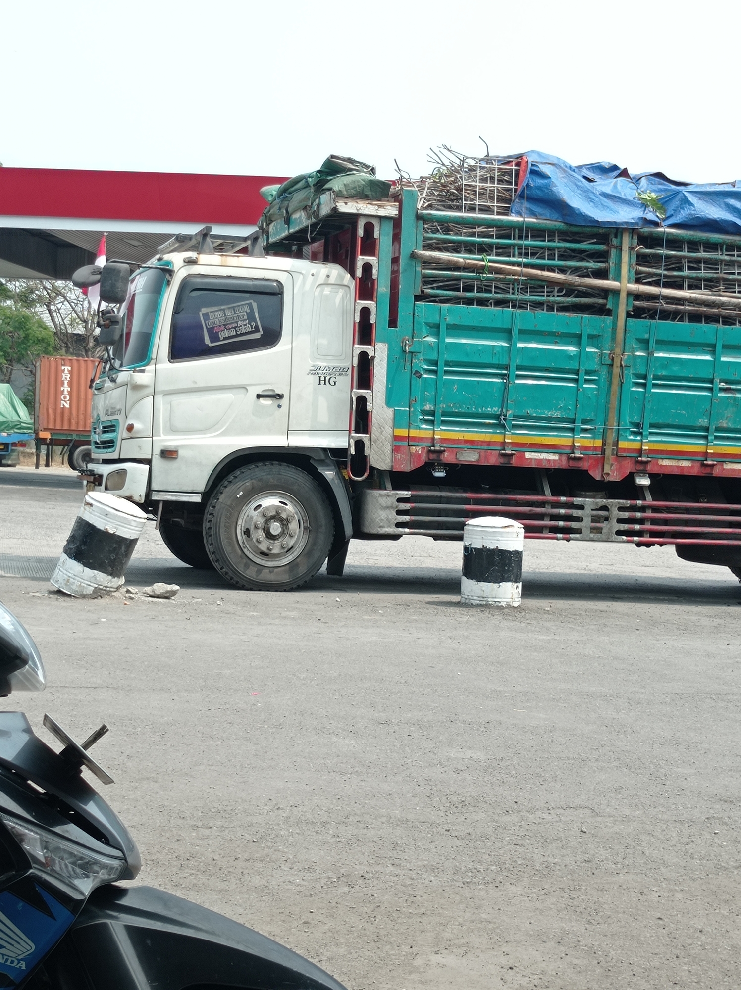 In Kramat, Indonesia, a large, teal-colored truck with a white cab is parked on an asphalt road. The truck is loaded with a bundle of wooden branches and is covered with a blue tarp. A sticker is affixed to the driver's side window of the truck, which reads "SUDAH LAMA" in larger font, and below it, "KITA JALAN BERSAMA" and "TIDAK ADA YANG SALAH?" in smaller font. In front of the truck, two white bollards with black stripes are positioned on the road; one is upright, and the other is tilted. Behind the truck, a red and white structure, possibly part of a building or canopy, is visible. The sky is overcast and white. The lower part of the image shows the front of a black motorcycle.