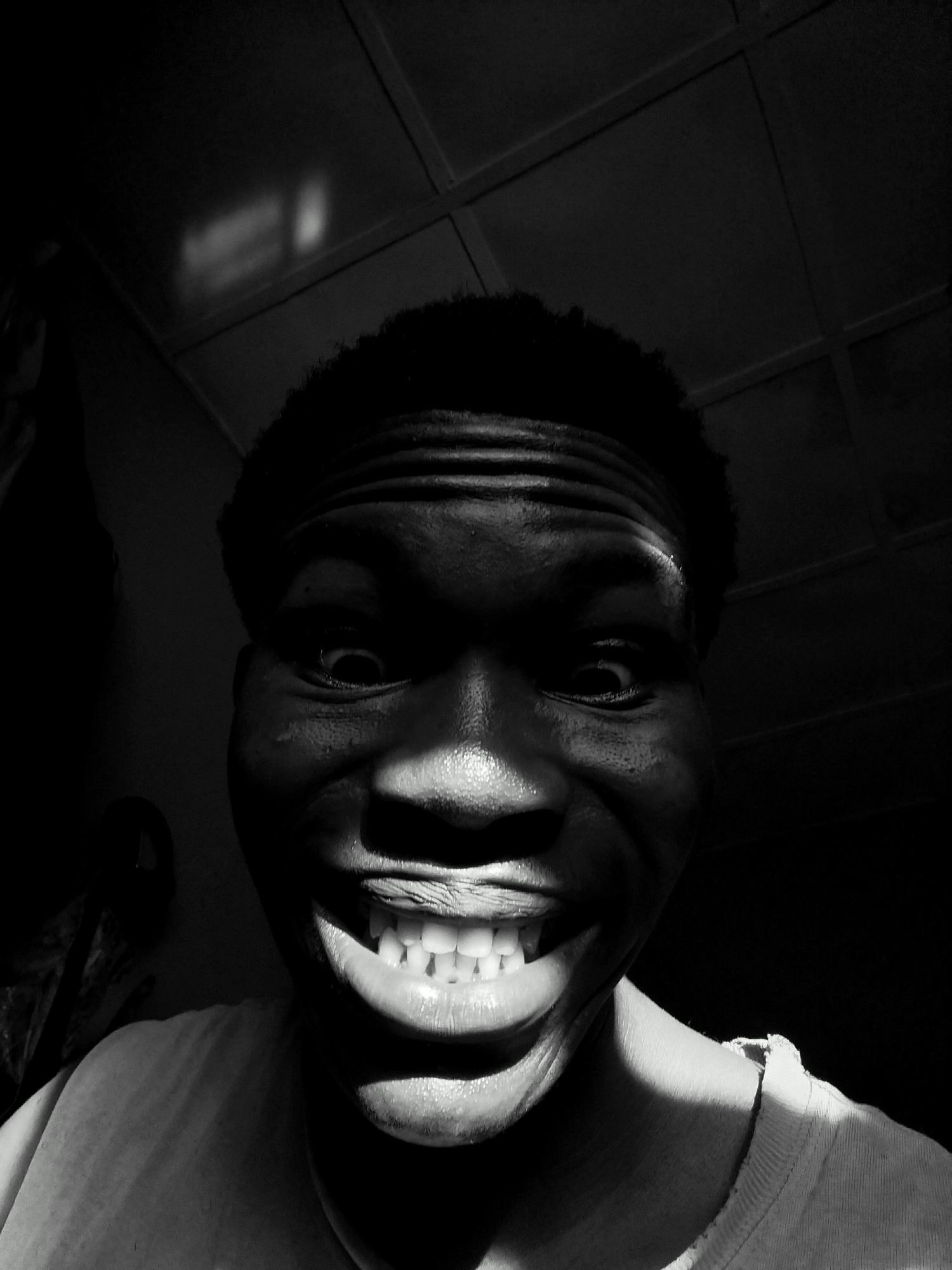 This striking black and white close-up portrait features a young individual, likely male, with dark skin and short, curly hair, captured in an indoor setting in Keffi, Nigeria. The subject looks directly at the camera with an intense and wide-eyed gaze, accompanied by a very broad, open-mouthed grin that reveals a full set of bright teeth. This expression conveys strong emotion, possibly extreme happiness, excitement, or a playful, mischievous demeanor.
The lighting is highly dramatic, characterized by a stark chiaroscuro effect. A strong, directional light source illuminates parts of the forehead, the bridge of the nose, the tops of the cheeks, and the teeth, creating bright highlights. Conversely, significant portions of the face, including the eyes, chin, and much of the background, are cast in deep, dark shadows, enhancing the contrast and adding to the intensity of the portrait. The subject is wearing a light-colored t-shirt, visible in the lower part of the frame.
The background is dark and indistinct, though faint outlines of what appear to be ceiling panels or beams can be seen in the upper corners, indicating an interior space. No discernible text is visible in the image. The high contrast and exaggerated facial expression make this a very impactful and memorable image.