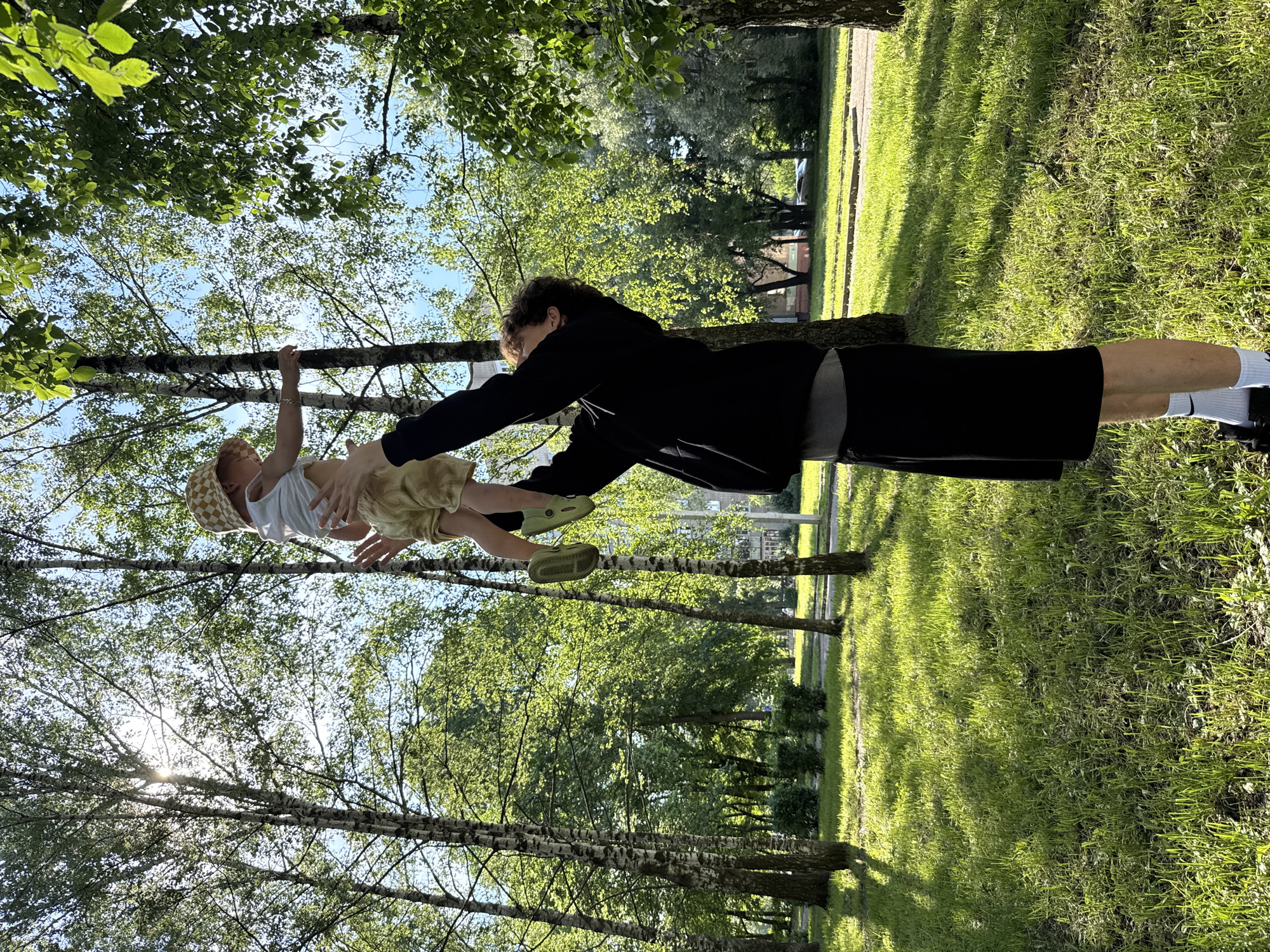 A young man is holding a baby in the air in an outdoor park. The man is wearing a black hoodie, grey t-shirt, and black shorts with white socks. The baby is wearing a white shirt, light yellow shorts, a checkered yellow hat, and green shoes. The background consists of green grass, trees with white bark, and a bright sky. Sunlight streams through the trees, casting shadows on the grass. In the distance, buildings and cars are visible.