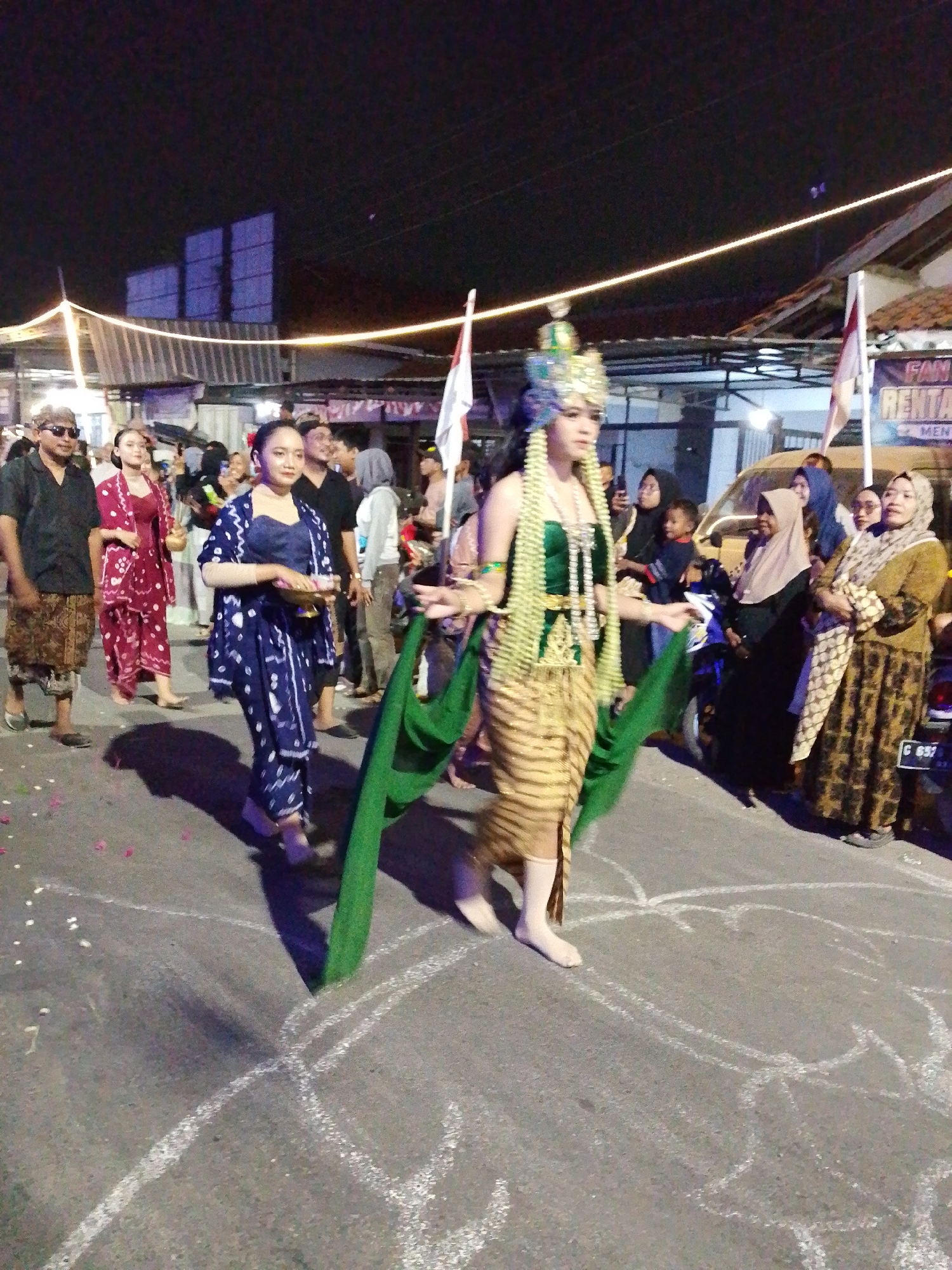 This image captures a nighttime street scene in Pagongan, Indonesia, likely during a cultural event or parade. In the foreground, a young woman in traditional attire is the central focus. She wears an elaborate headdress, a green beaded necklace, and a gold and beige striped skirt, with a flowing green fabric draped over her arms. Her feet are bare, and she is walking on a street marked with white chalk drawings.
To her left, a woman in a dark blue patterned outfit with a matching jacket and pants is walking, holding a decorative bowl. Next to her, another woman is dressed in a maroon patterned outfit. Several other individuals, men and women, are visible in the background, some wearing headscarves and traditional Indonesian clothing. They appear to be spectators or participants in the event.
The environment is dimly lit by string lights, creating a festive atmosphere. Buildings and stalls line the street, and a vehicle is partially visible on the right side of the image. Flags are also present, with one on the right displaying text that includes "RENTAL." The ground is paved asphalt, and there are scattered flower petals on the surface. The overall impression is one of a lively community gathering celebrating local traditions.