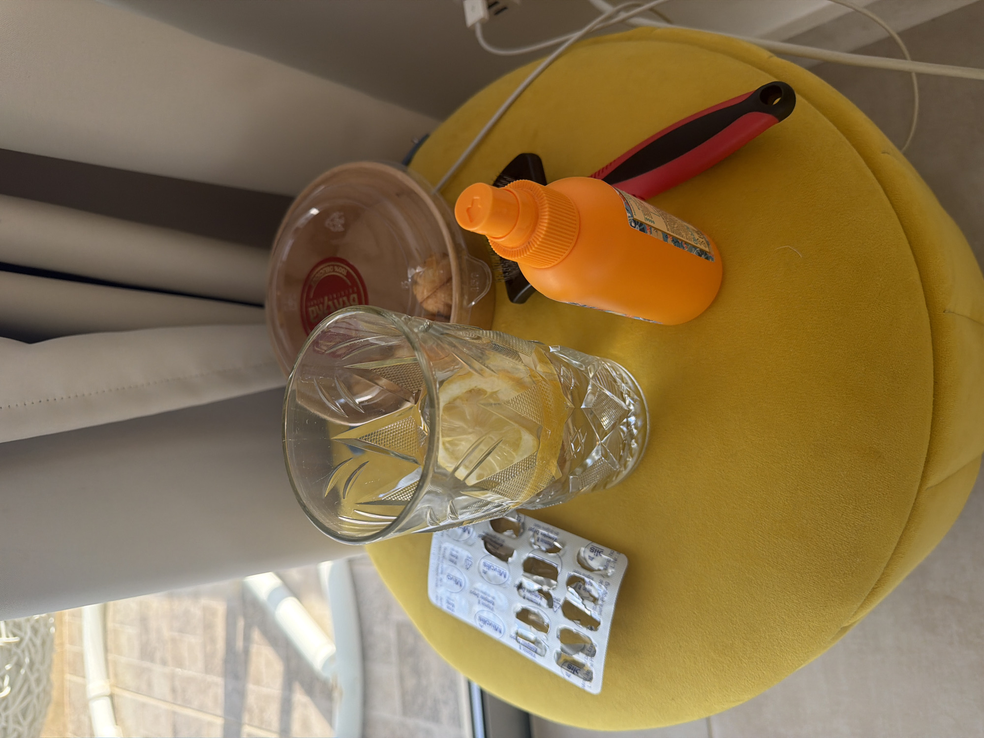 An outdoor scene in Peyia, Cyprus, features a round, yellow ottoman with various items on its surface. To the left, a blister pack of opened medication is visible. Next to it, a clear glass contains water and a slice of lemon. To the right of the glass, an orange spray bottle with a black and red handle lies on its side. Behind these items, a clear plastic container with a red label and some food items is partially obscured. The background includes a window with vertical blinds and a white electrical cord running across the ottoman. The ground appears to be tiled.