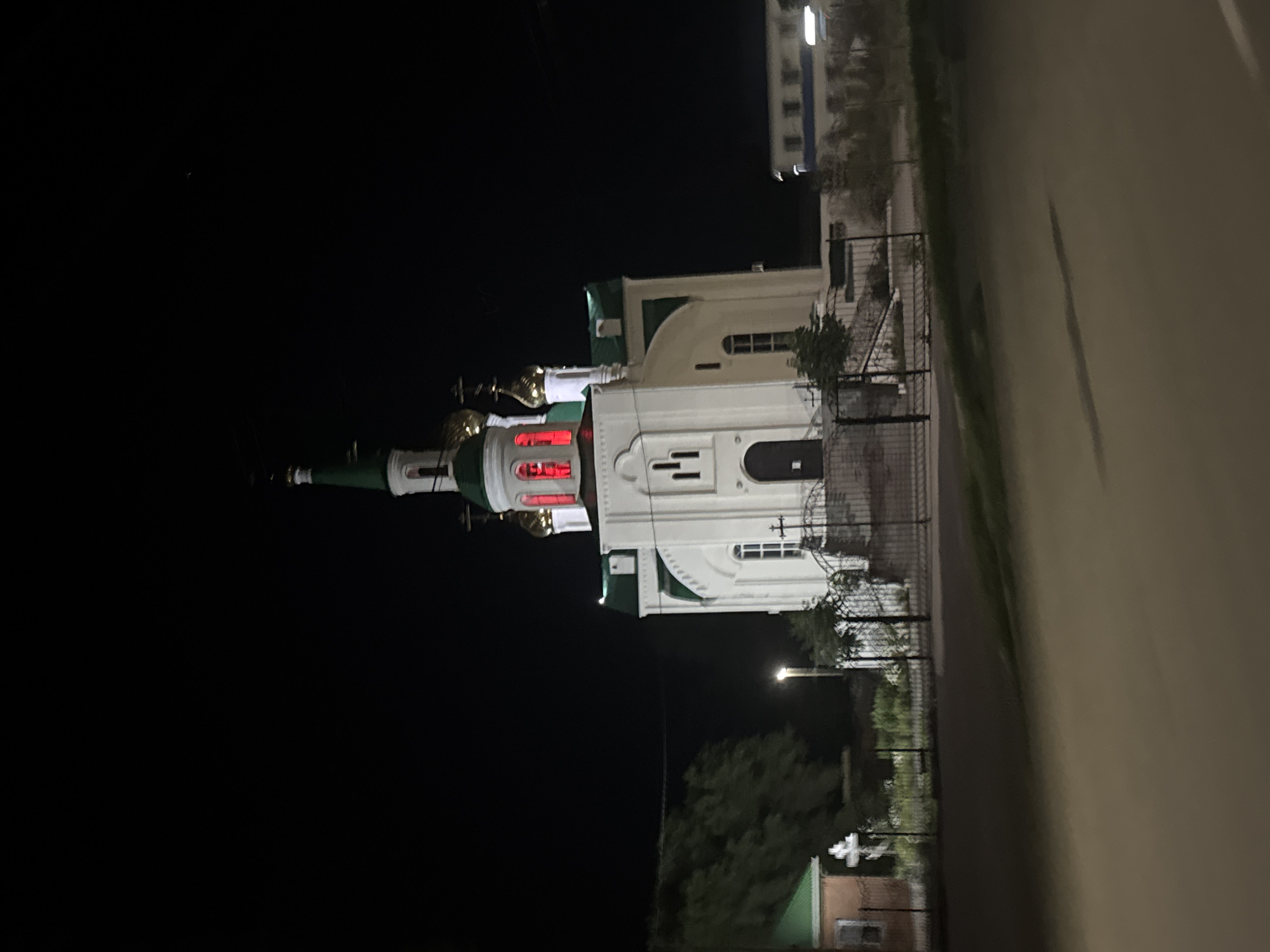 The image shows a church at night in Yegorlykskaya, Russia. The church has a white facade with green accents and a gold dome. Red lights are visible inside one of the church's windows. The scene is dark, indicating it is nighttime. A road is visible to the right of the church, and some trees and a fence are also present. There is also a building in the background. The overall mood is peaceful. No text is visible in the image.