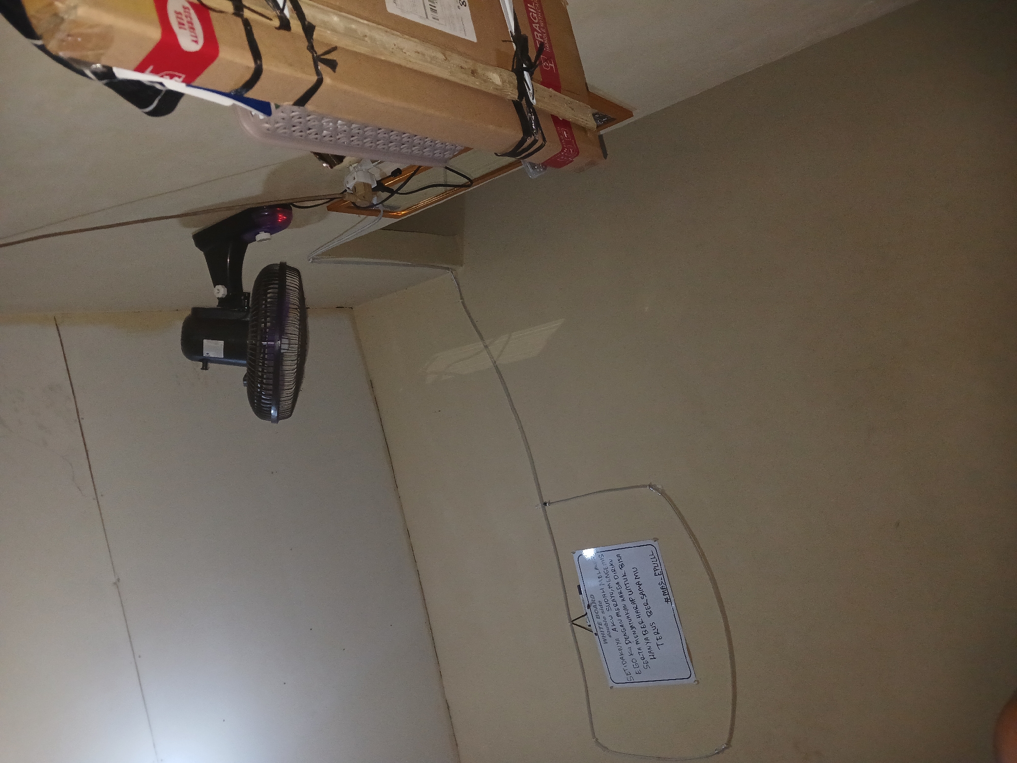 The image shows an indoor scene with a white ceiling and light brown walls. A black oscillating fan is mounted on the wall to the right of center. To the right of the fan, a stack of cardboard boxes is secured to a wooden shelf that is also attached to the wall. The boxes have red and white warning labels, one of which reads "FRAGILE HANDLE WITH CARE". On the wall to the left of center, a small whiteboard with the words "WHITE BOARD double side" is hanging. Below this, handwritten text in Indonesian can be seen. The text appears to be a message or a poem. The overall lighting is dim, with a bright light source visible at the top left of the image. The image appears to be taken from a low angle. The location is identified as Cikedokan, Indonesia.