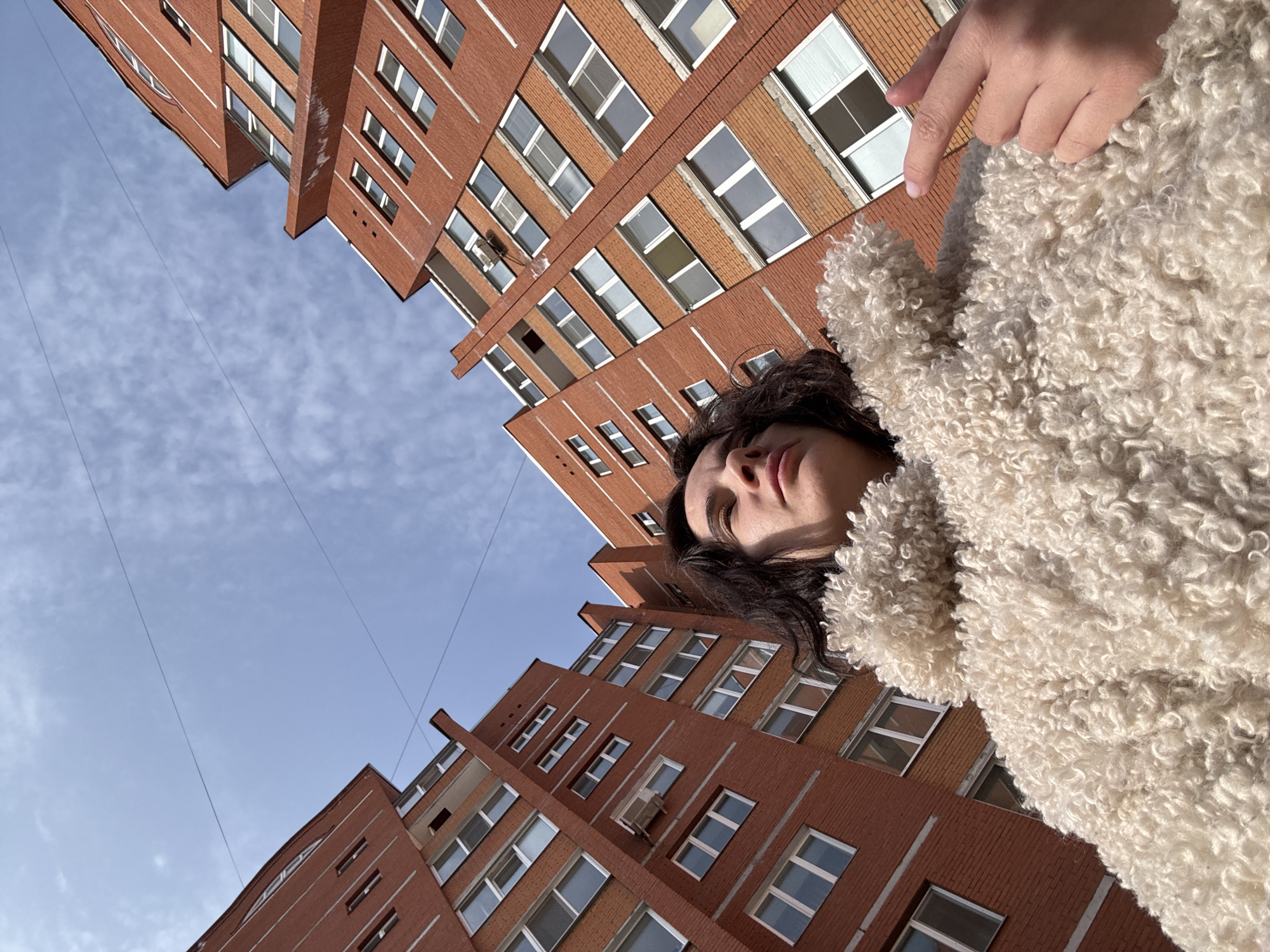 This image, taken in Chaikovsky, Russia, presents a low-angle view of an individual against a backdrop of urban architecture and sky. A person, whose head and upper torso are visible, occupies the foreground. The individual has dark, curly hair and is wearing a voluminous, light-colored, textured coat, likely made of faux fur or fleece. Their face is turned slightly to the right, with eyes gazing upwards. The right hand is partially visible in the lower right quadrant, with the index finger extended.
The background consists of two tall, multi-story apartment buildings constructed from reddish-orange brick. These buildings feature numerous rectangular windows with white frames and white vertical lines painted on their facades. An external air conditioning unit is mounted on one of the buildings. Above the structures, the sky is clear and blue with scattered wispy white clouds. Two thin, dark utility wires traverse the upper sky. The scene is brightly illuminated by direct sunlight.