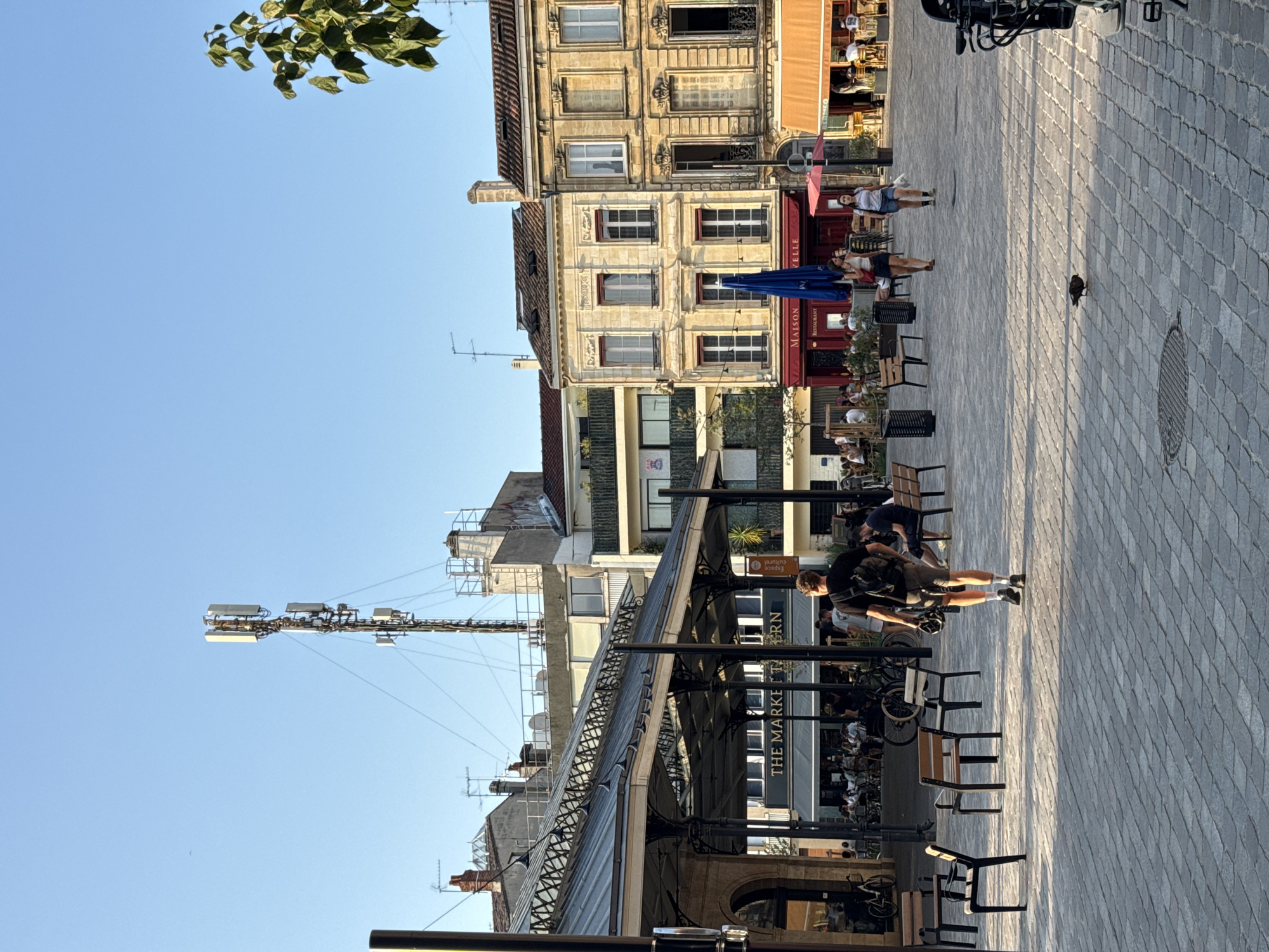 The image depicts an outdoor market square in Bordeaux, France, under a clear blue sky. The foreground is paved with grey cobblestones, and several empty chairs and benches are scattered around. A man with a backpack is walking away from the camera, holding a helmet in his right hand. In the background, there is a covered market structure with the words "THE MARKET TAVERN" visible on its facade. Beyond that, several multi-story buildings with traditional European architecture line the square, some housing cafes and restaurants with outdoor seating. A prominent cell tower stands tall in the distance, and a small bird is visible on the cobblestone ground in the mid-ground. The overall atmosphere is relaxed, suggesting a daytime scene in a public urban space.