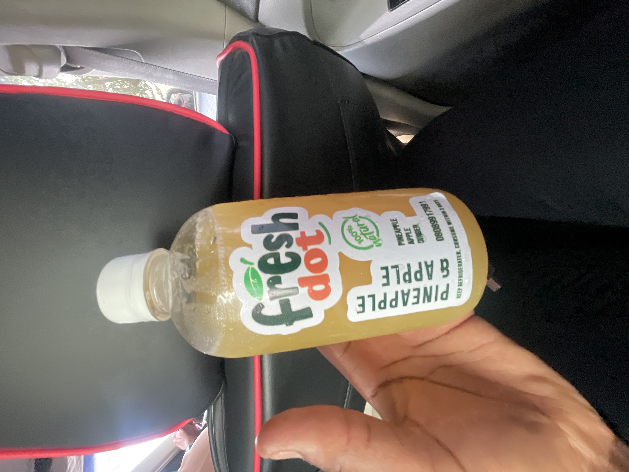 A hand holds a bottle of "Fresh Dot Pineapple & Apple" juice inside a car. The bottle has a white cap and a label with green and red lettering. The car's interior features black seats with red trim and grey dashboard elements. The image's setting is likely a vehicle's interior in Abuja, Nigeria.