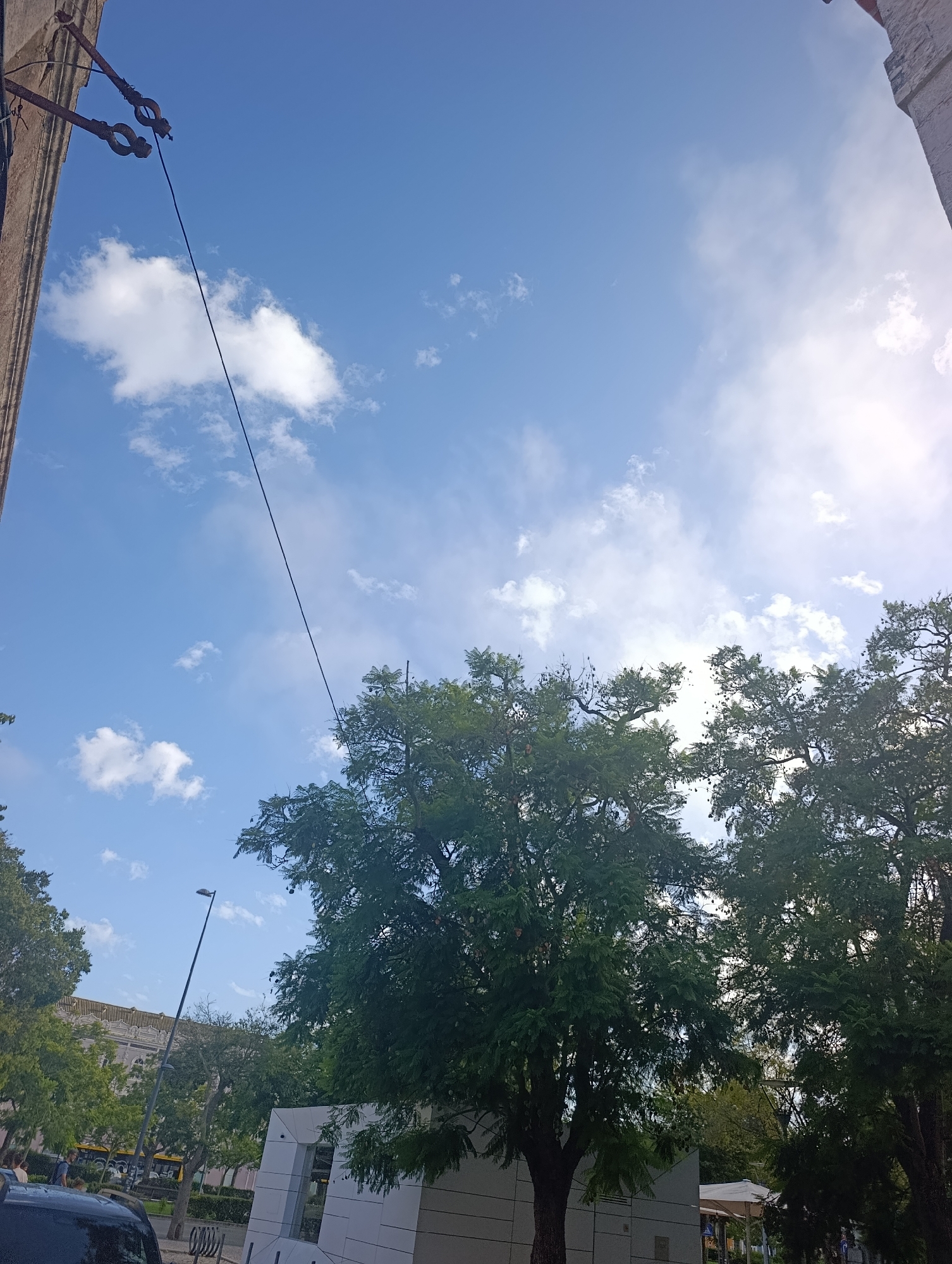 The image captures a street view in Setúbal, Portugal, under a bright blue sky dotted with scattered white clouds. On the left side, a portion of a building's facade is visible, with what appears to be exposed wiring attached to a support structure. A dark-colored vehicle is parked in the foreground. Beyond the vehicle, a street with a lamppost and trees lines the area. In the background, several trees with dense green foliage dominate the scene, partially obscuring a building with a yellow-tiled roof and a modern-looking white structure. A few individuals can be observed walking on the sidewalk and near the bus.