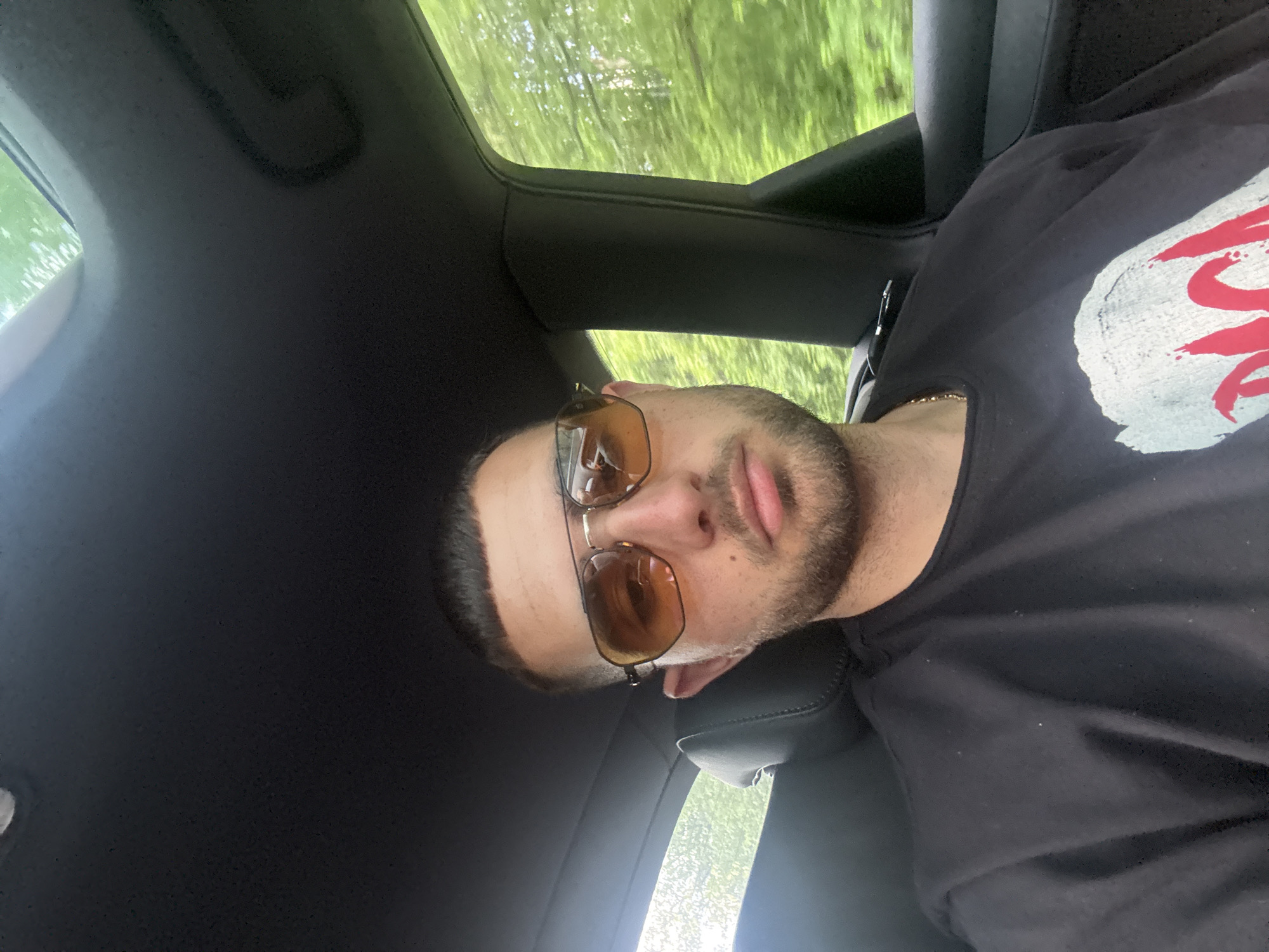 A man wearing sunglasses and a black t-shirt with a white and red design is in the driver's seat of a car. Trees are visible through the sunroof. The interior of the car is black. The location is likely a road in Rahovtsi, Bulgaria based on previous context.