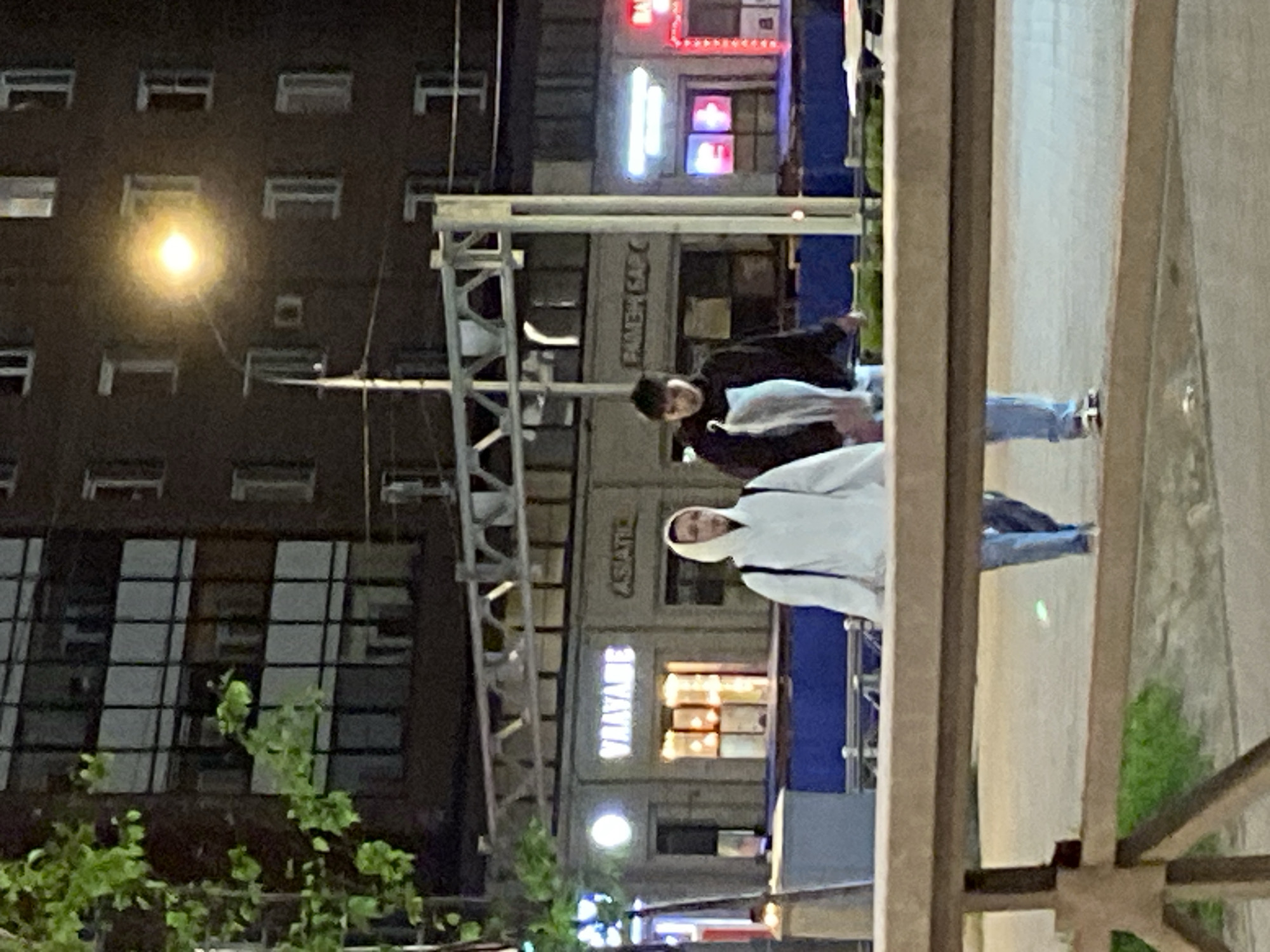 The image shows two people walking down a street at night. One person is wearing a white hoodie and the other is wearing a black jacket. They appear to be near a building with a sign that says "BAYANE". The location is likely Saint Petersburg, Russia. The scene is viewed through a structure, possibly a fence.