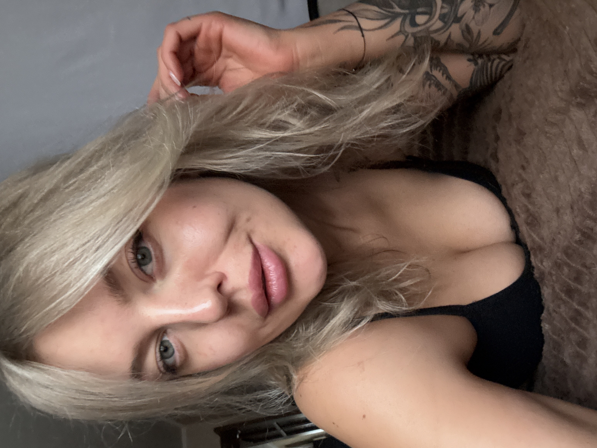 A woman with light blonde hair is lying down. Her head is tilted to the side, and she is looking at the camera. She has fair skin, and her lips are pink. She is wearing a black top. Her left arm is raised, and there are tattoos visible on her arm and shoulder. She is lying on a brown, furry surface. The background is out of focus and appears to be a light grey wall. There is no indication of the location being Borzya, Russia.