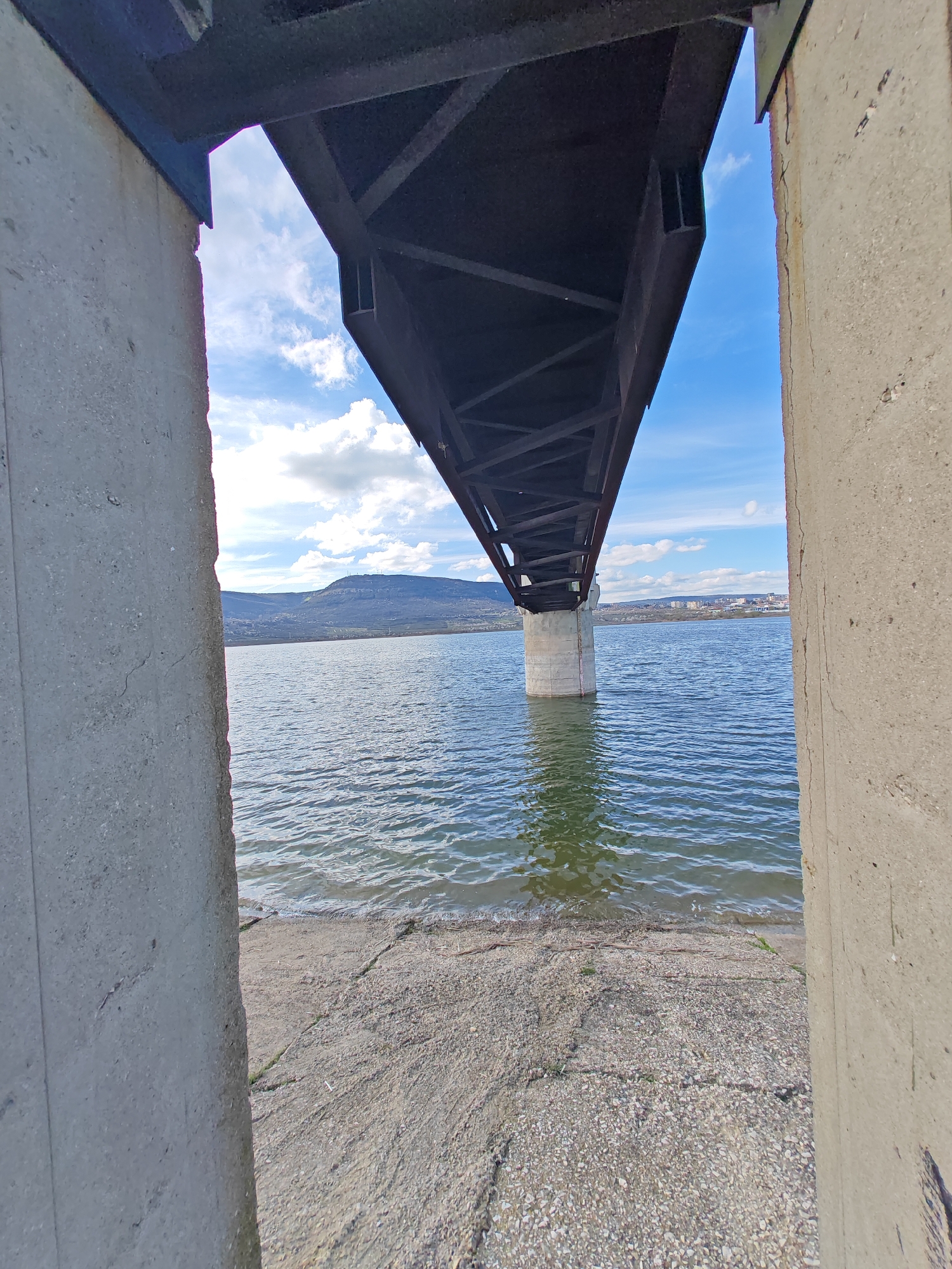 The image shows the underside of the Asparuhov Bridge in Shumen, Bulgaria, as seen from a low vantage point between two concrete pillars. The bridge's steel structure is dark and contrasts with the bright sky visible between its supports. Water covers the scene, with a shoreline where the water meets a concrete or stone surface. The water appears calm, with small ripples. In the background, a distant hill or mountain range is visible beneath a partly cloudy sky. No people are visible in the image. The focus is on the structural elements of the bridge and its interaction with the surrounding environment.