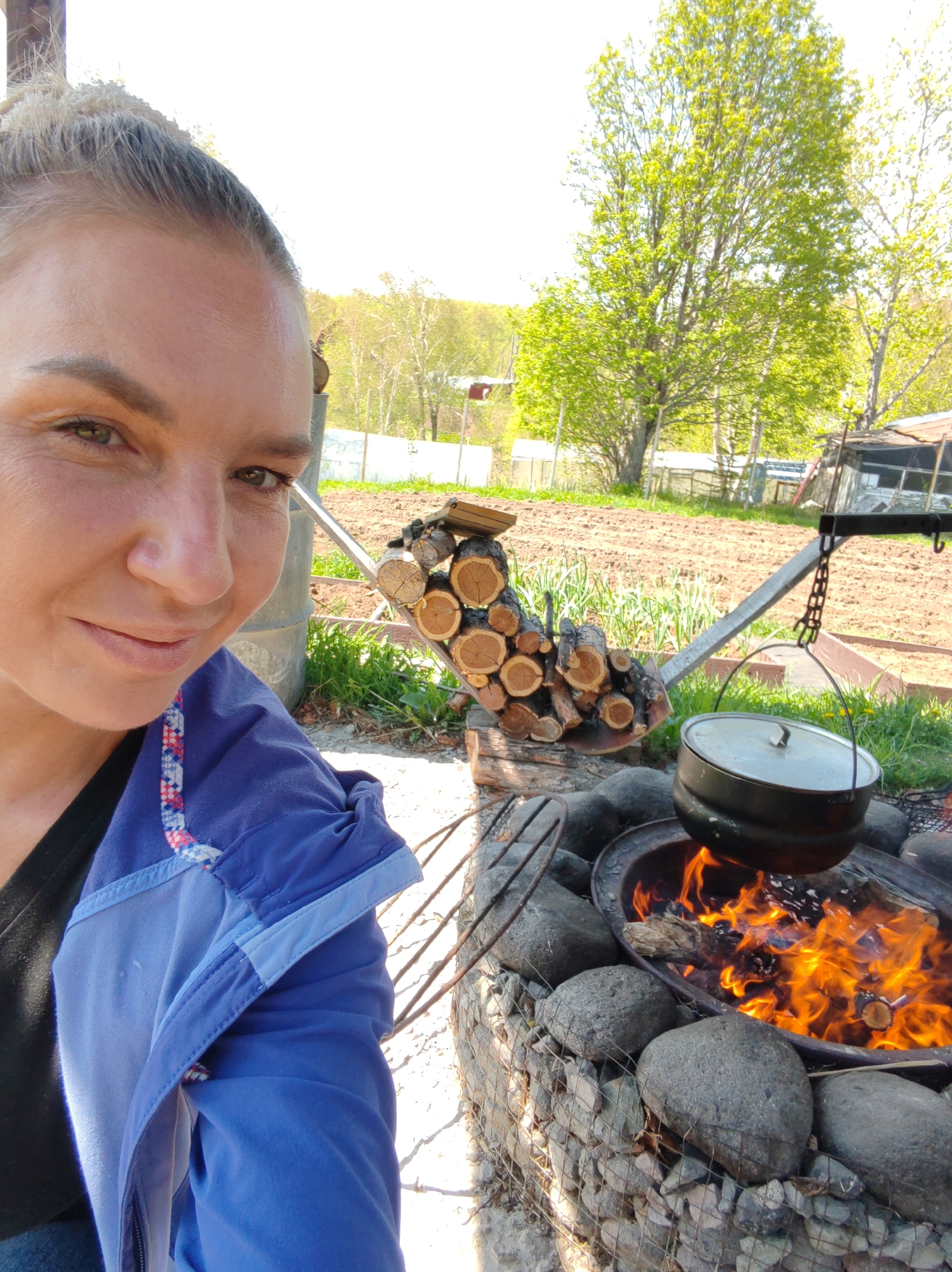 An adult female captures a close-up selfie in an outdoor, rural setting. She has light skin, hair pulled back into a bun on top of her head, light brown/blonde eyebrows, and light-colored eyes. She exhibits a slight smile and looks directly at the camera. She wears a blue jacket with lighter blue trim details on the collar and cuffs, and a visible red, white, and blue braided drawstring or trim inside the collar, over a dark-colored shirt.
To her right and slightly behind her, an outdoor cooking setup is active. A circular fire pit is constructed from large, grey, irregular stones contained within a metal mesh cage. Inside the pit, a dark metal basin holds an active fire, characterized by bright orange and yellow flames consuming small logs. Suspended directly over the flames is a dark-colored, possibly cast-iron, cooking pot with a metallic lid and a wire bail handle. The pot hangs from a metal chain, which is attached to a rustic, dark metal support frame, partially visible. Adjacent to the fire pit, a neat stack of cut firewood logs, displaying concentric rings and some darker, charred ends, rests on a wooden support.
The background reveals a rural landscape under a clear, bright sky. A large expanse of tilled, brown earth with distinct linear rows and emerging green sprouts suggests a garden or agricultural field. A prominent, tall, deciduous tree with vibrant green spring foliage stands in the mid-background. Other trees and sparse green vegetation are scattered throughout the scene. A white fence line runs horizontally across the mid-ground. In the further distance, several small structures are faintly visible, including what appears to be a greenhouse or shed on the left, and a weathered wooden building with windows on the far right. The scene is brightly illuminated by natural daylight.
The location is Unknown, Unknown.