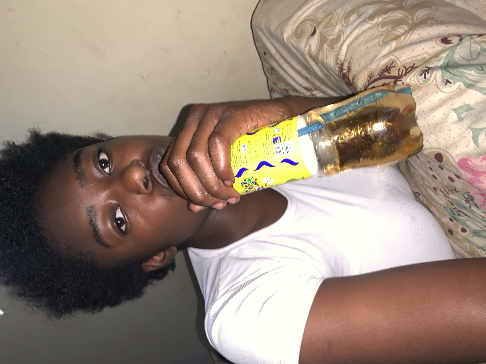 A person with dark skin and short, curly, dark hair is angled towards the camera. They are wearing a white ribbed t-shirt and are holding a yellow juice box with a blue straw in their left hand to their mouth. The box is partially transparent and contains a light brown liquid. The person is lying on a bed with a patterned comforter in the background. The location is New Stadium, Nigeria.