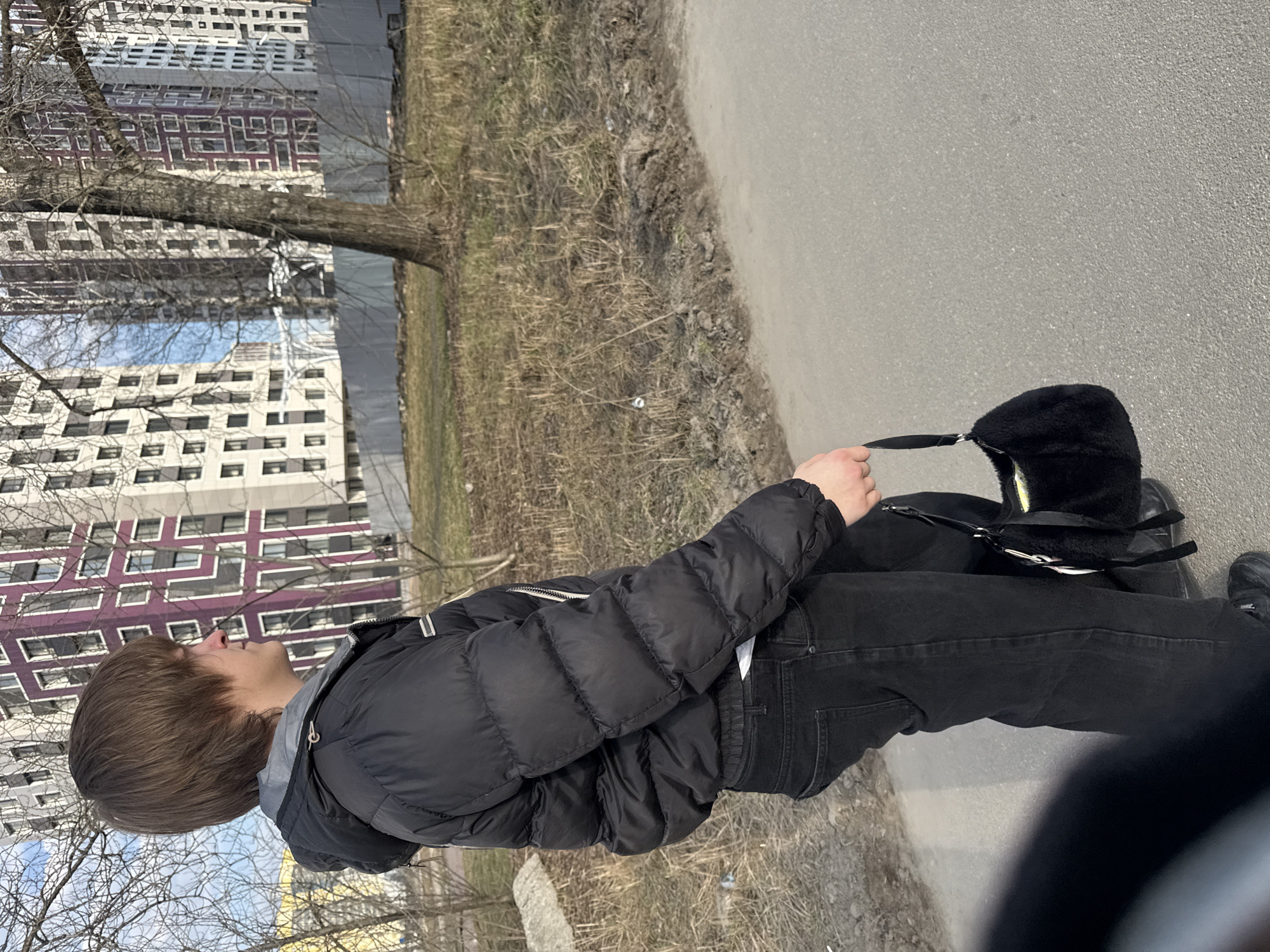 A young person, possibly male, is walking a dog on a leash alongside a road or pathway. The person is wearing a black puffer jacket, black jeans, and dark shoes. The dog appears to be wearing a dark carrier or backpack. The surrounding environment includes buildings in the background that are typical for Saint Petersburg, Russia, bare trees, and patches of grass and dirt along the side of the path. The weather appears to be sunny with a bright sky.