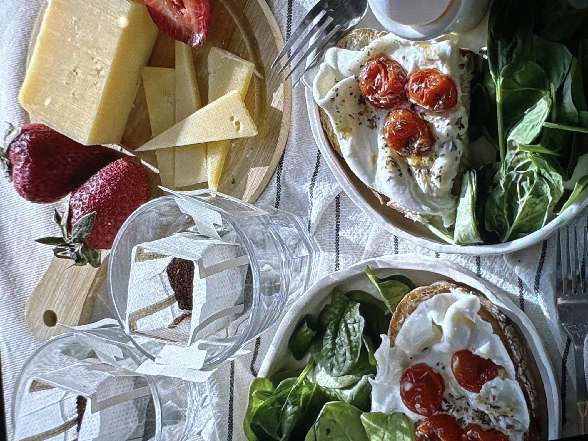 The image depicts a breakfast or brunch setting. On the left, there's a wooden board with cheese and strawberries. Nearby are transparent glasses filled with what appears to be coffee filters or tea bags. Two plates hold toast topped with white cream, roasted tomatoes, and fresh spinach. Silver forks are visible near the plates. The scene is set on a striped tablecloth. There is no information available in the image about the location being in Yerevan, Armenia, or any other city. The image depicts a meal.