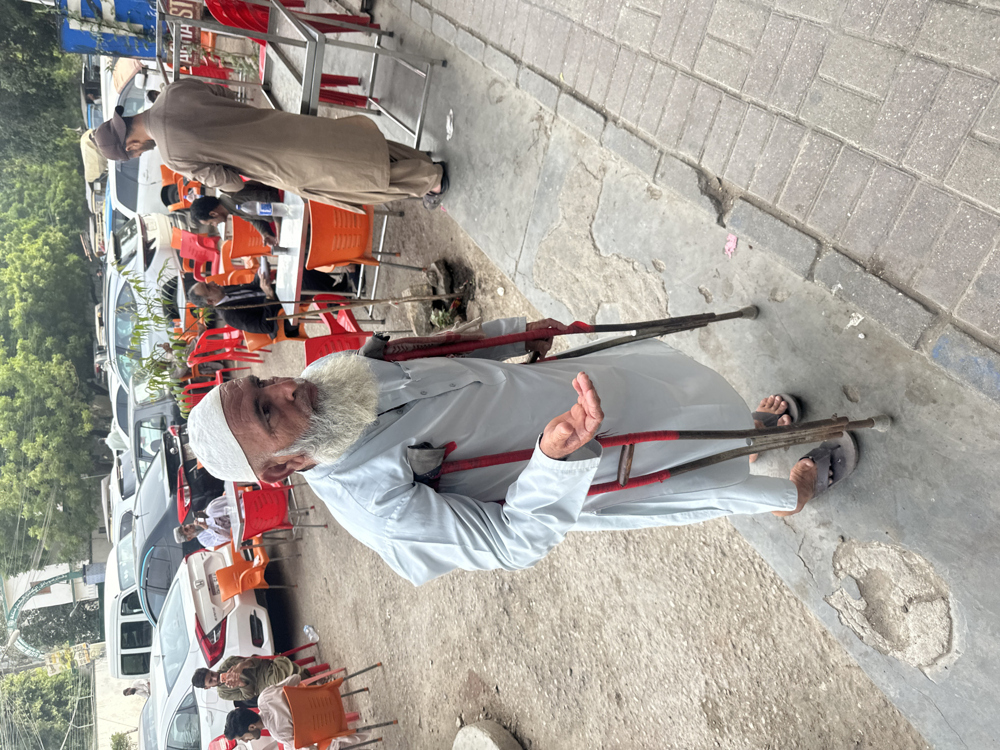 A full shot captures an elderly man, with a white beard and matching cap, standing outdoors with the assistance of two red and silver crutches. He is dressed in a light blue shalwar kameez. His left arm is bent at the elbow as he holds the crutches, with his hand slightly open, palm up, in a gesture that could indicate asking for something. The background suggests an urban environment in Karachi Division, Pakistan, with various cars parked behind him. Several people are visible sitting at tables in the background, some on orange and red chairs, suggesting an outdoor seating area, possibly a cafe or restaurant. The ground in the foreground is concrete, with some cracks and patches, transitioning to a brick-paved sidewalk to the right. Trees and buildings are visible in the distance.