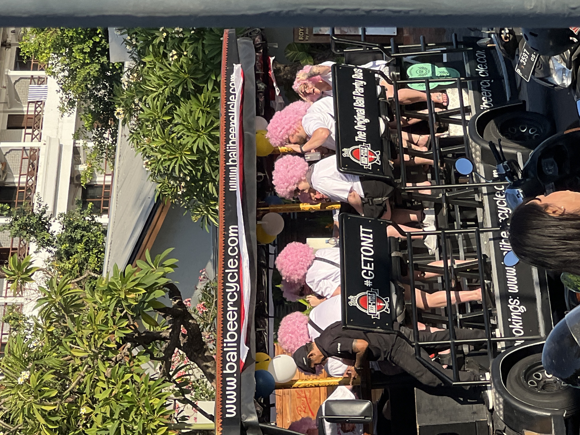 In Kuta, Indonesia, a group of individuals on a "Bali Beer Cycle" are captured in an outdoor setting. The Beer Cycle, a pedal-powered vehicle designed for group tours, features a canopy with the website "www.balibeercycle.com" displayed prominently. Several people are seated on the vehicle, most wearing bright pink wigs, suggesting a celebratory or themed event. One person in the foreground, wearing a black cap and black shirt, appears to be operating or serving on the vehicle. Others are seen holding drinks and interacting. The vehicle itself has branding that reads "Bali Beer-Cycle" and "#GETONIT", with additional text "The Original Bali Party Bus". The background shows lush greenery and a building with balconies, consistent with a tropical urban environment. The scene is well-lit, indicating daytime conditions.