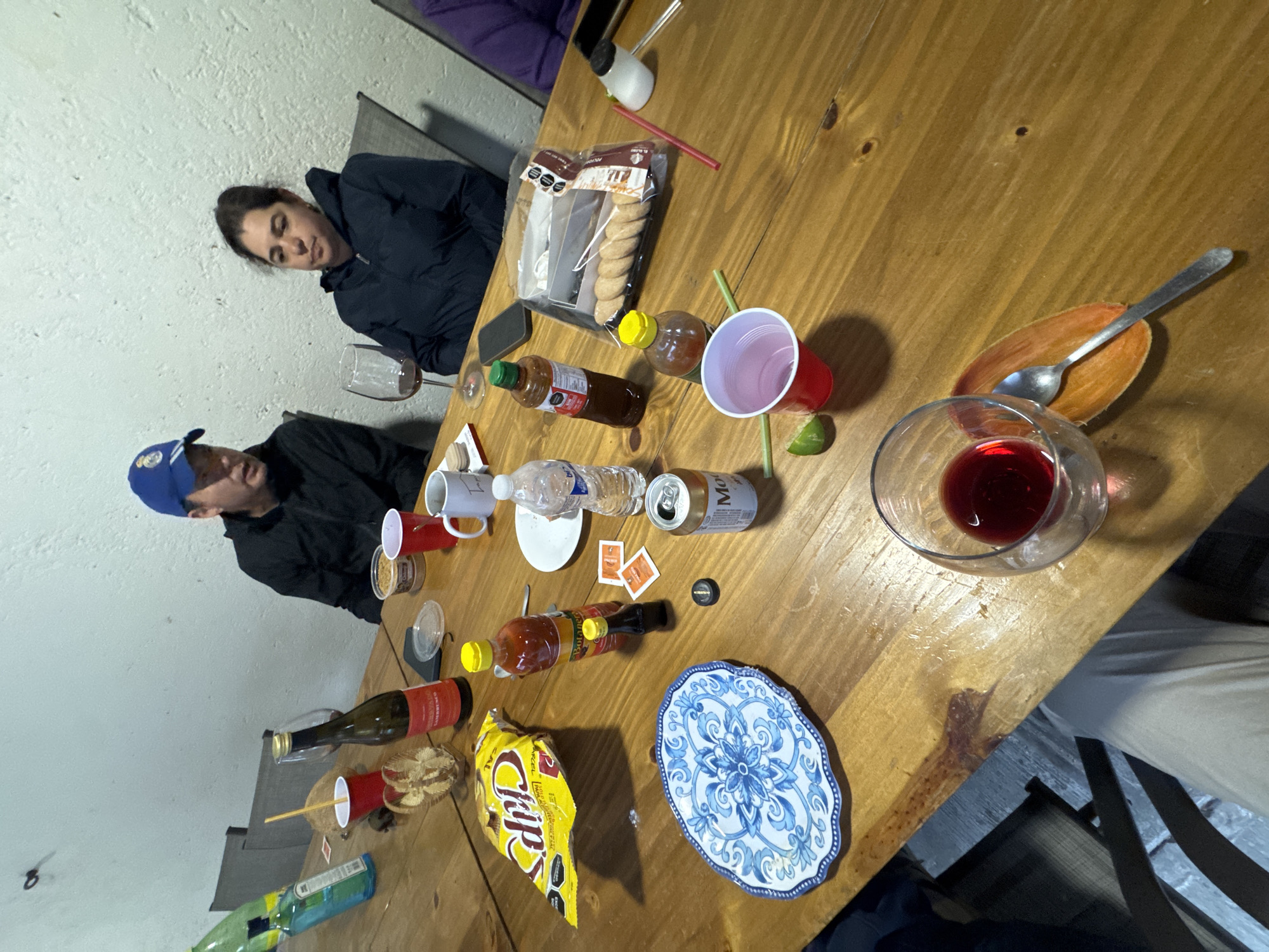 **Scene:**
A cluttered, slightly haphazard gathering around a long, light-colored wooden table.
**People:**
Two individuals are partially visible. One person wears a black jacket and a blue baseball cap. The other person wears a black jacket.
**Objects:**
The table is covered with assorted food and drink items, including:
* Several bottles of hot sauce.
* Bottles of alcohol.
* Red Solo cups.
* A water bottle.
* A can of beer.
* A bag of potato chips.
* A package of cookies.
* Wine glasses, one containing red wine.
* A plate with a lime.
* A spoon on a plate.
* A smartphone.
* Salt packets.
**Environment:**
The location appears to be an indoor space with a white painted wall and simple furniture, possibly in a residential setting.
**Location:**
The specific location in the city of Puebla, Mexico is not identifiable from the scene.
