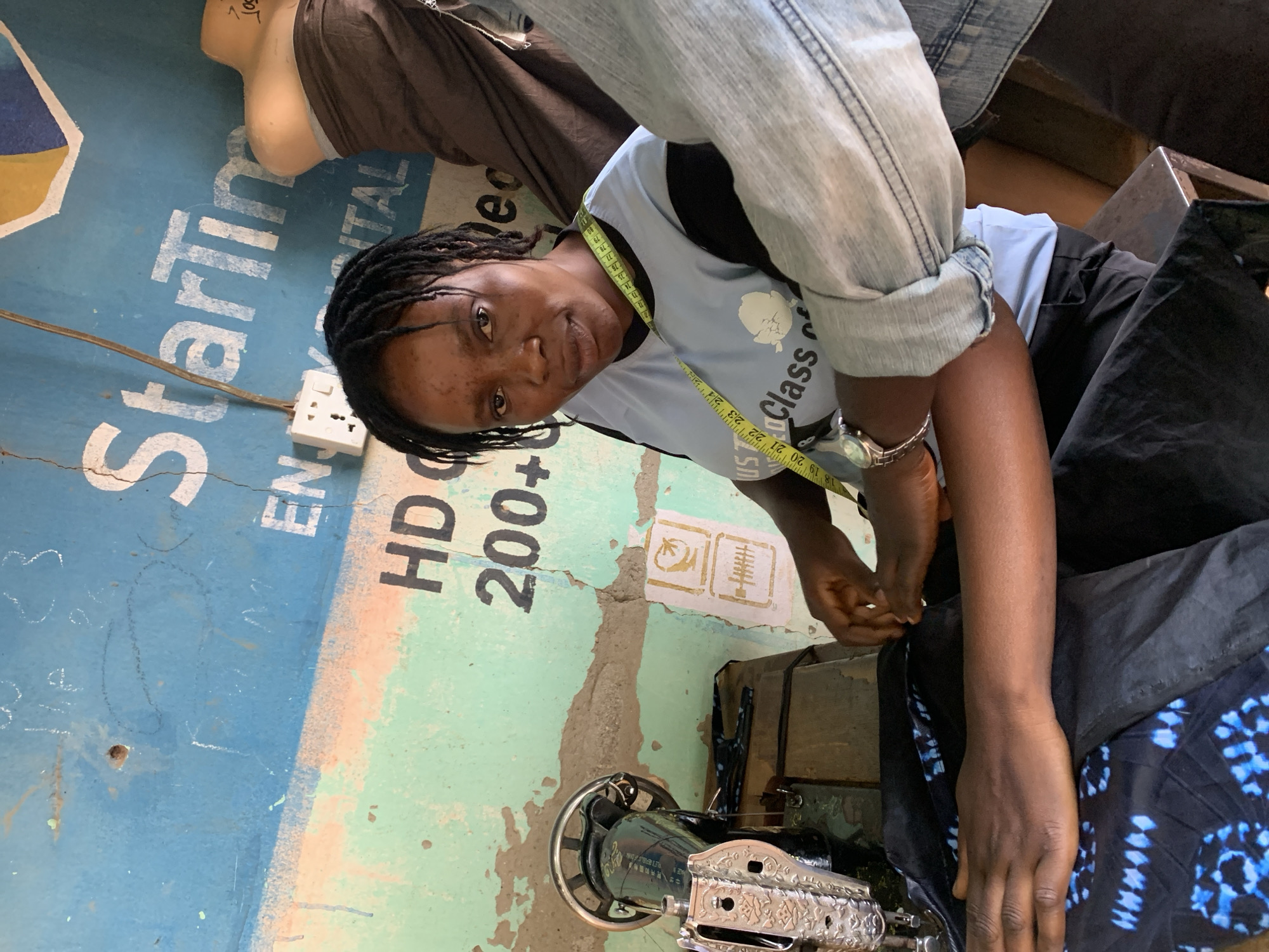 A medium shot depicts a woman with braided hair and a tape measure around her neck, seated at a vintage sewing machine. She is wearing a light blue t-shirt with a graphic, and her hands are positioned as if she is preparing to sew a piece of dark blue patterned fabric. The sewing machine is a metallic color with decorative details. Behind the woman is a faded wall painted in blue and green, with the words "StarTin," "ENTAL," "HD C," and "200+" visible, indicating a sign. The presence of a mannequin and clothing suggests the location of the image is Maigizo, Nigeria.