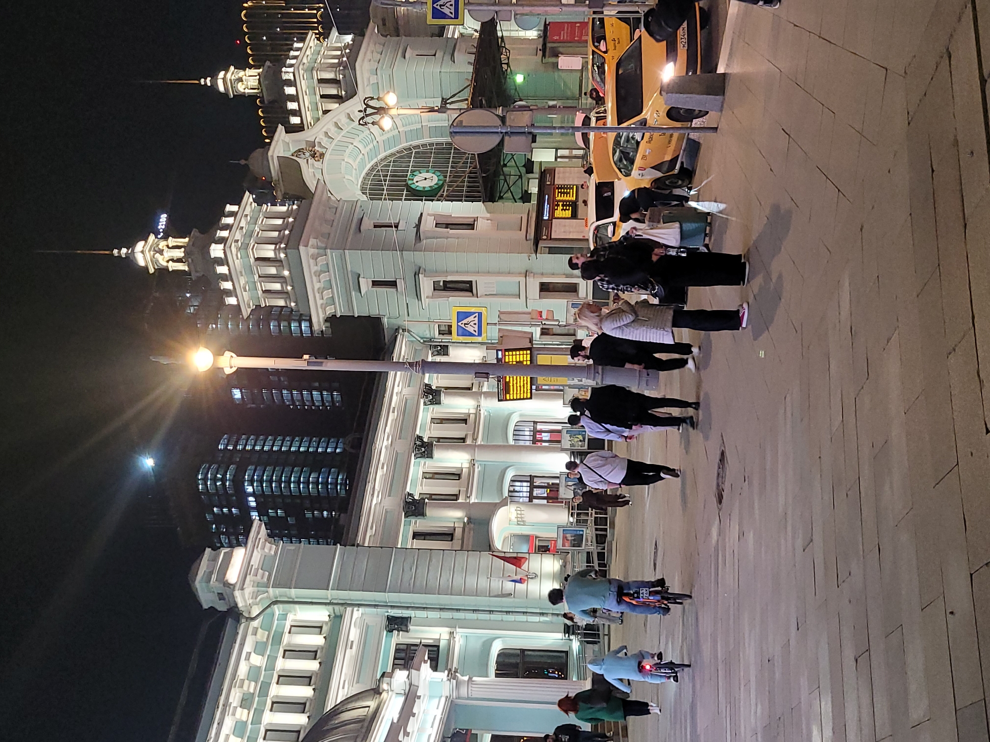 At night, a crowded sidewalk in Moscow, Russia, fronts the Kazansky train station, where people are standing, walking, or riding bicycles. Several yellow taxis are parked along the curb. The station's illuminated clock face reads approximately 9:15. A modern skyscraper is visible in the background to the left of the station. Pedestrian crossing signs are present. The ground is paved with light-colored stone tiles.