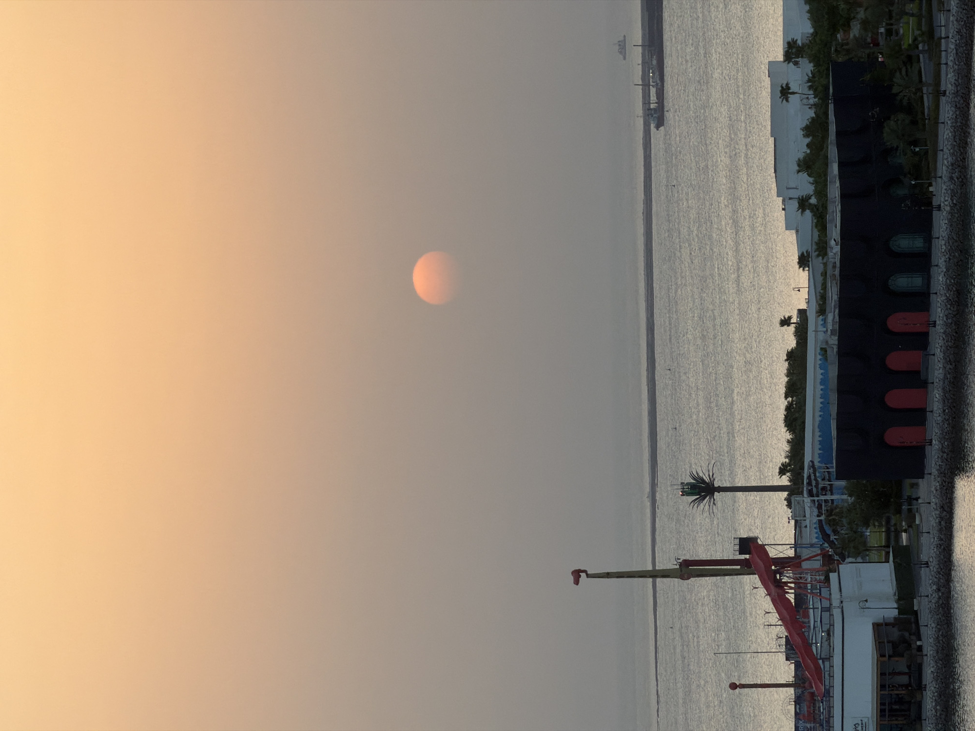 The image depicts a wide shot of a waterfront cityscape during what appears to be either sunrise or sunset. The sky is a hazy gradient of peach and orange, with the sun a prominent, diffused orange orb in the upper right quadrant. Below the sky, a calm body of water, likely the sea, stretches horizontally.
In the foreground, a paved area runs along the edge of the water, with a stone embankment. Several buildings are visible, with a predominantly white structure on the right and a dark building with arched red and green doorways in the center. To the left, part of a white building with a shaded outdoor seating area is visible. Behind these, a palm tree stands tall with a cluster of lights on top.
Further back and to the left, elements of an amusement park are present, including a tall, red-and-yellow structure with a red slide. In the water, a large cargo ship is seen in the distance on the right, and a smaller boat is closer to the foreground. Another similar structure to the red slide amusement park ride is visible on the far left. The overall atmosphere is serene, with the soft lighting creating a hazy, ethereal effect.