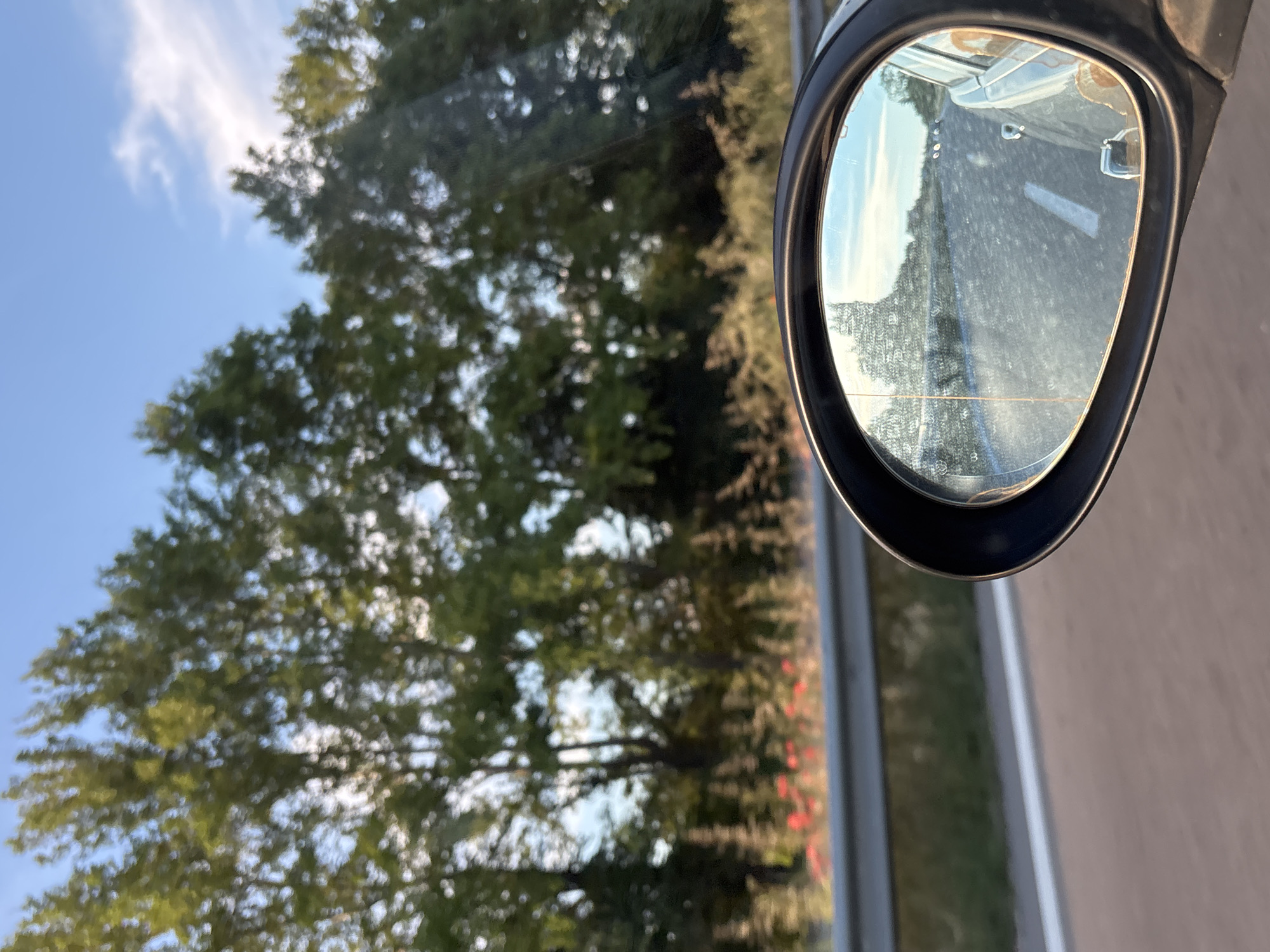 The image shows a reflection in a car's side mirror, depicting a road, trees, and the sky. The reflection shows a road with a white line marking the lane, and a silver car is also visible in the reflection, suggesting the viewer is traveling in a car. Beyond the road, there are trees with green foliage, and the sky is blue with some light clouds. The environment appears to be natural, possibly a rural area or a highway passing through a green landscape. There are no people or specific urban elements visible in the reflection or the surrounding environment. The location is identified as Ignatievo, Bulgaria.