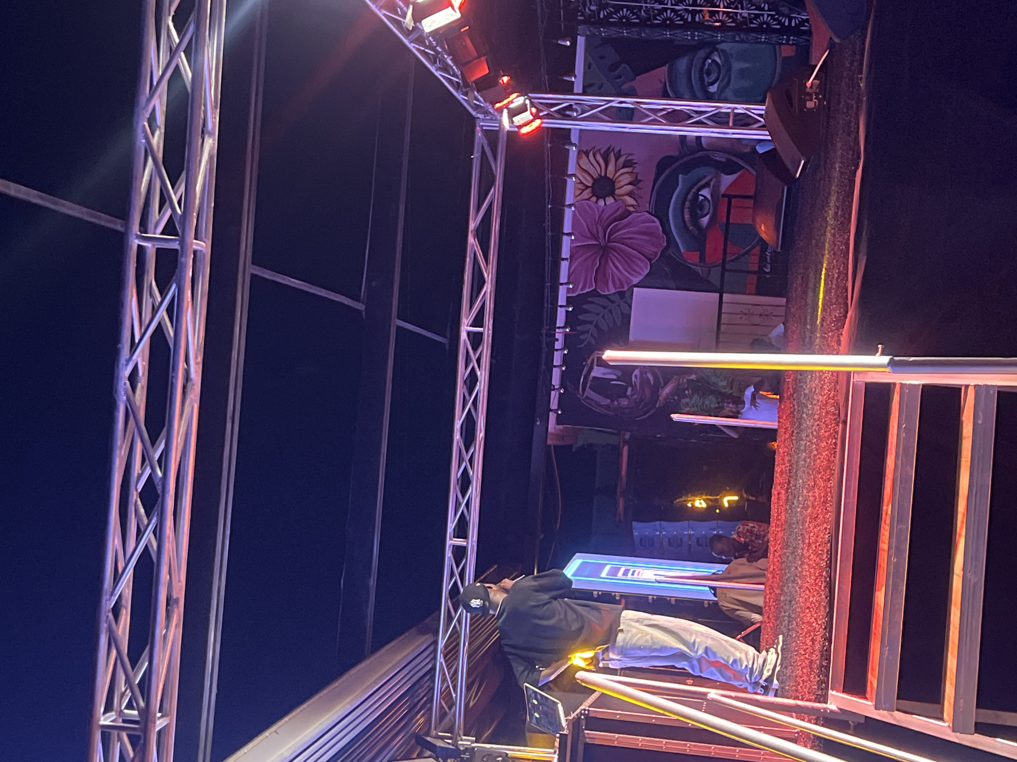A low-angle shot depicts an outdoor performance stage located in Howayi, Nigeria. The stage is constructed of silver metal scaffolding, with red stage lighting attached. The backdrop is a painted mural featuring flowers and stylized eyes. A man wearing a black jacket and a baseball cap stands near the edge of the stage, operating electronic equipment. Behind the stage, buildings and streetlights are visible in the distance. A red barrier is positioned along the stage's edge.