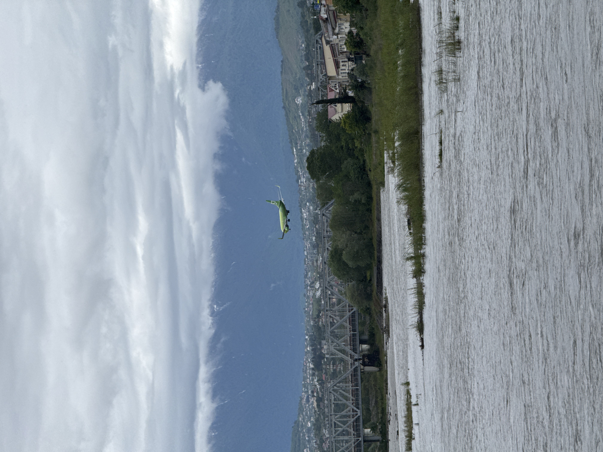 A landscape image shows a river flowing along the right side. On the left side of the river, vegetation lines the river's edge. A bridge crosses the river in the middle of the image, connecting the foreground to the background. In the background, a city lies against a mountain range. An airplane flies in the sky against the backdrop of the mountains and city. The sky is filled with heavy clouds. The image is taken vertically. The location appears to be Sochi, Russia.