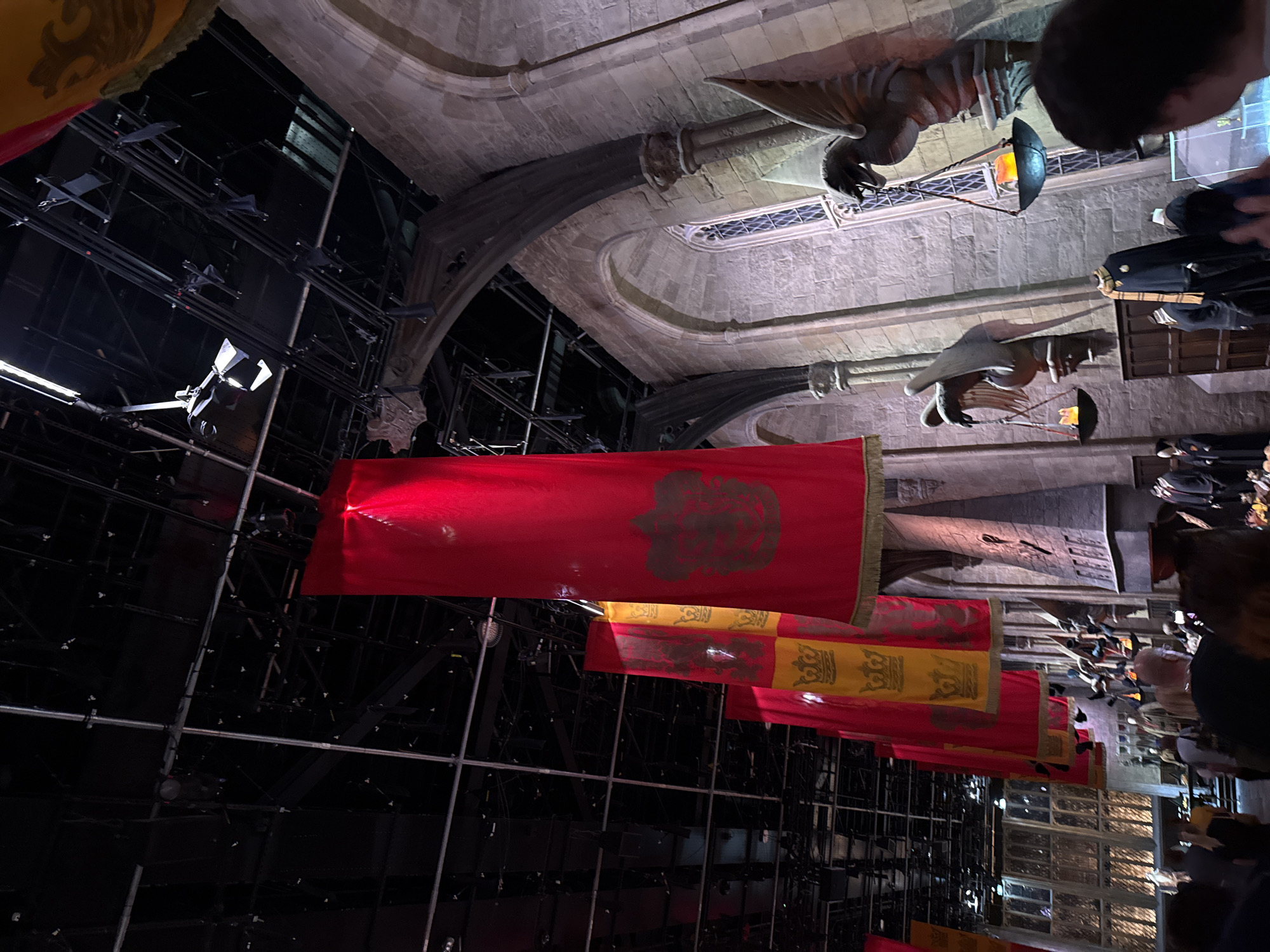 The image depicts the Great Hall set from Warner Bros. Studio Tour London - The Making of Harry Potter, located in Leavesden, near Watford, United Kingdom. The scene features iconic red and gold banners with house crests hanging from a high, industrial-looking ceiling with exposed lighting rigs. Stone archways and walls, characteristic of a medieval hall, are visible on the right side, adorned with stone gargoyle-like figures holding lanterns with flickering faux flames. A long wooden table is set up in the background with various props and costumes, including Hogwarts robes, on display. Several people are present, observing the set from a distance, with some in the foreground looking towards the display. The lighting is dim and atmospheric, accentuating the grandeur and mystical ambiance of the Harry Potter universe.