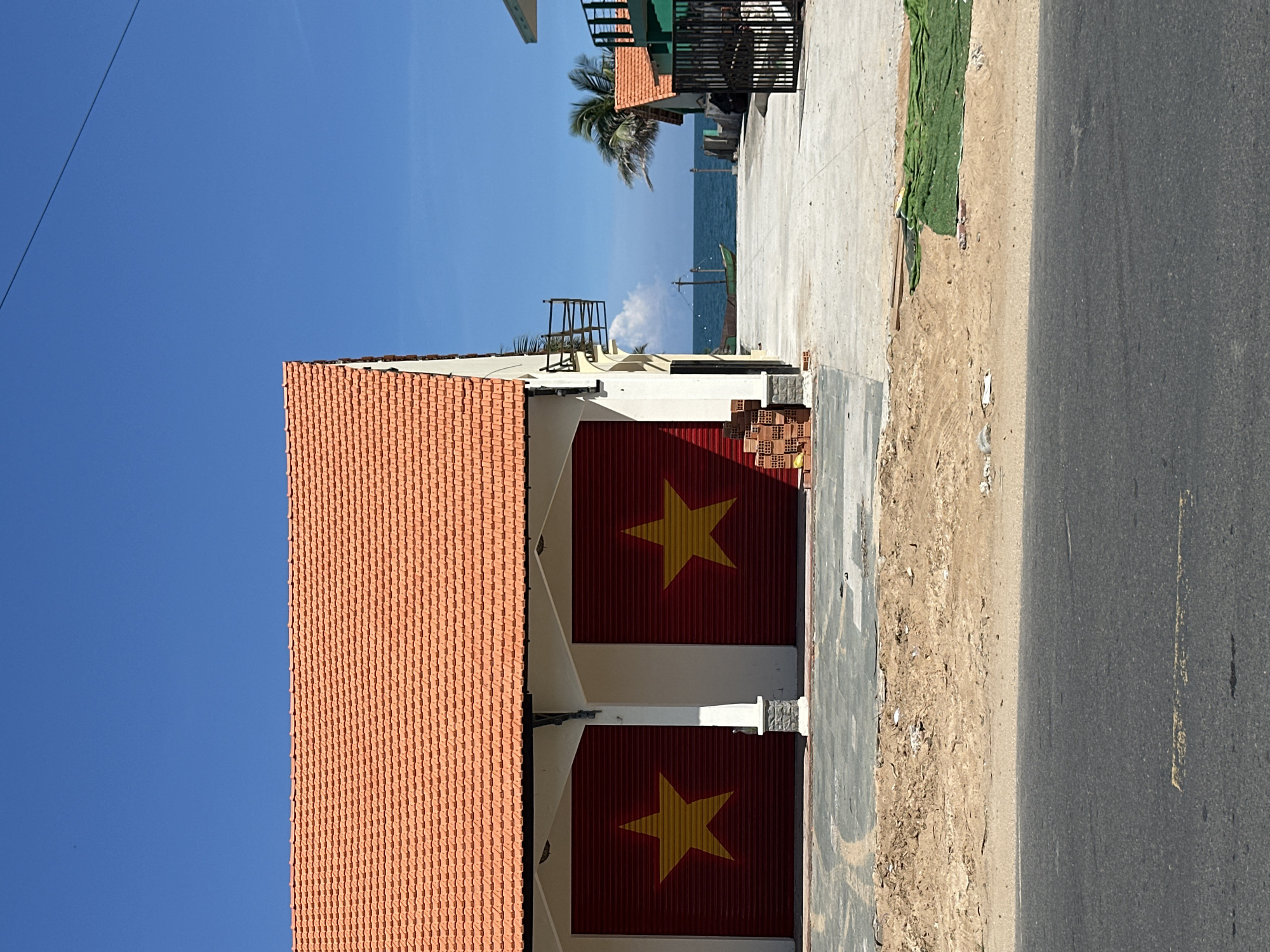 This is a medium shot, eye-level, outdoor, daytime photograph of a building in Phu Thuy Ward, Vietnam. The building is painted white and has an orange tiled roof. On the front of the building are two large red garage doors, each with a yellow star. To the left of the doors is a stack of bricks. In front of the building is a paved area, a patch of dirt, and a road. Beyond the building, the ocean can be seen, with a small boat on the water and a palm tree. The sky is clear and blue.
