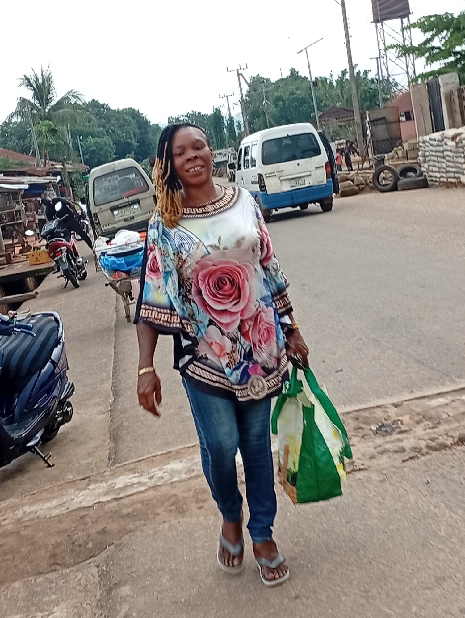 A full shot shows a woman walking along a paved street in Nsugbe, Nigeria, carrying a green and white shopping bag. She is wearing a colorful blouse with a floral print, blue jeans, and gray flip-flops. The woman has a gold bracelet on her left wrist and her hair is styled in blonde and dark braids. The street is lined with shops, parked motorcycles, and a white van. In the background, trees and a water tower are visible. The sky is overcast.